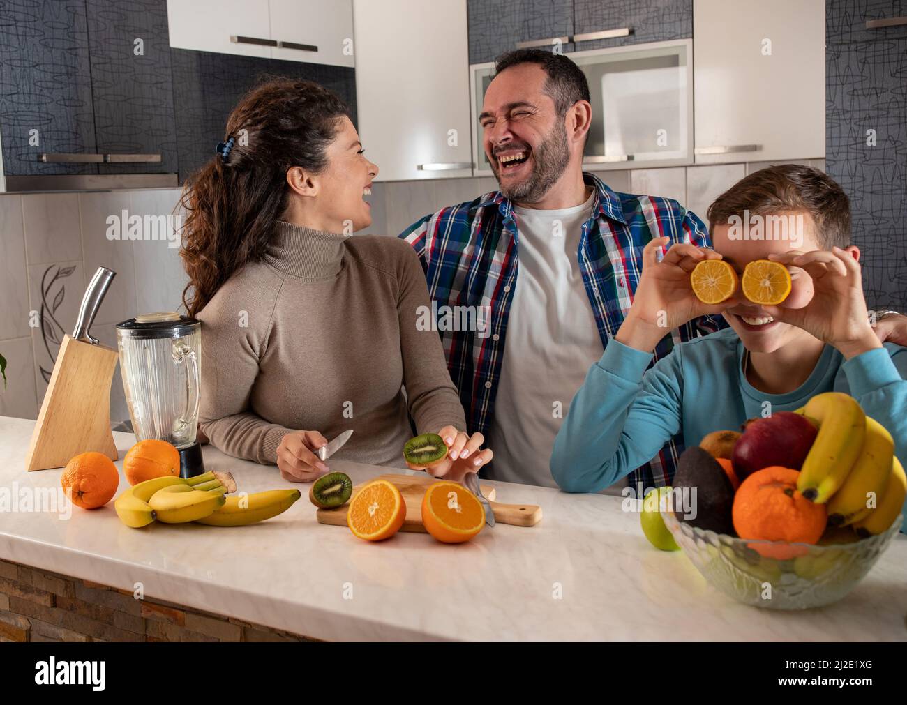 Madre e padre in piedi in cucina ridendo al loro figlio che sta ingannando intorno mentre tiene le arance sugli occhi Foto Stock