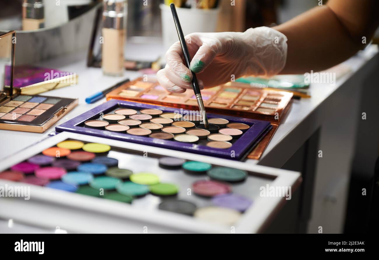 Make-up artista con cosmetici professionali in salone di bellezza. Primo piano della mano femminile in guanti sterili che tengono la spazzola cosmetica trucco sopra la tavolozza di ombretti. Foto Stock