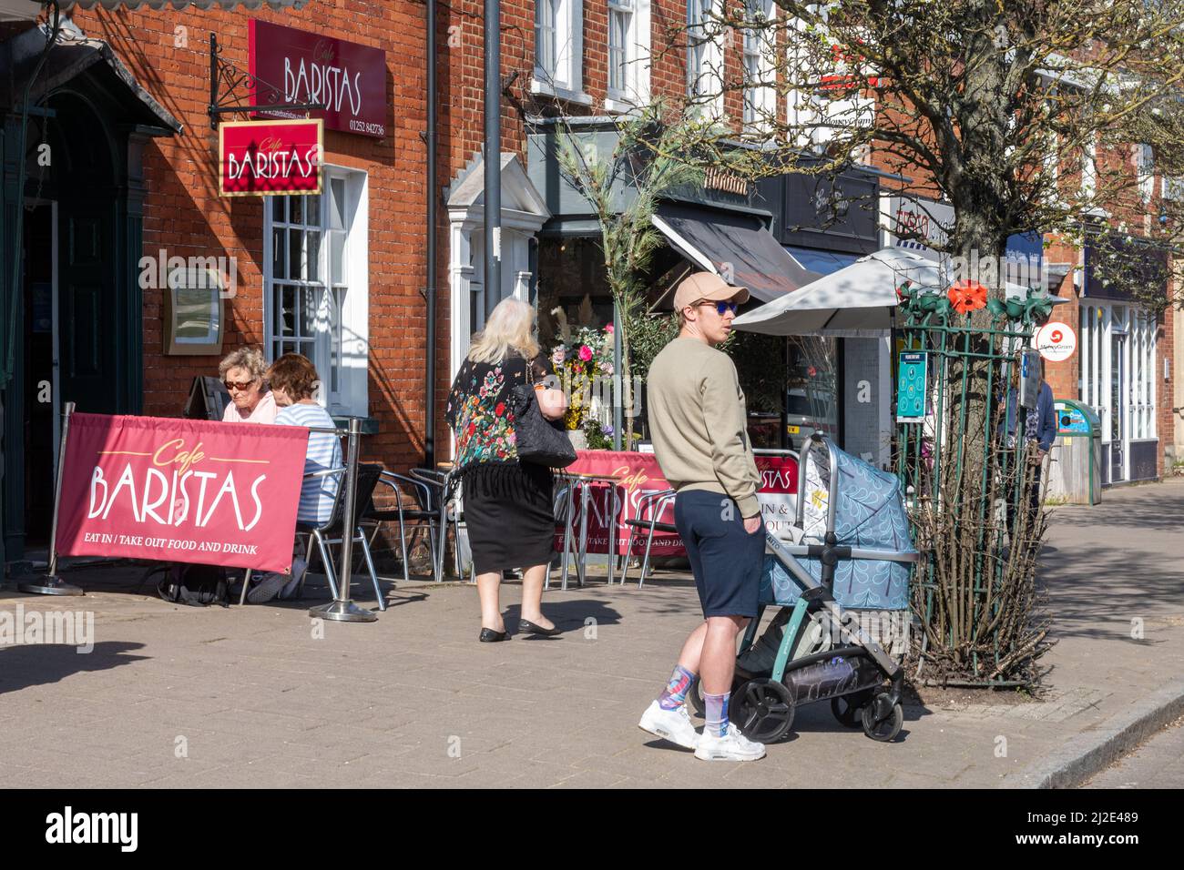 Hartley Wintney Village Center, Hampshire, Inghilterra, Regno Unito. Vista della High Street con il giovane uomo che si cura di un bambino in un gram Foto Stock