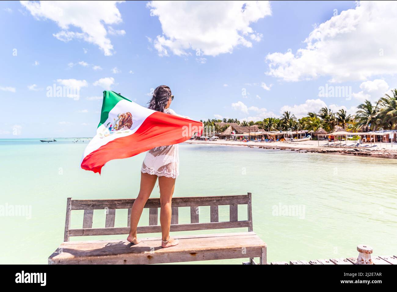 Donna si erge su una panca di legno che regge la bandiera messicana guardando verso il mare con spiaggia, palme e case. Foto Stock