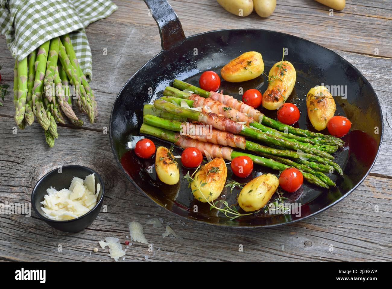 Asparagi verdi fritti avvolti in pancetta e serviti con patate alle erbe e pomodori con parmigiano grattugiato in una vecchia padella di ferro Foto Stock