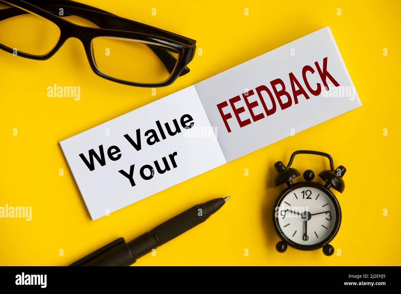 Testo su blocco note con occhiali da lettura, penna e sveglia su sfondo giallo - diamo valore al vostro feedback. Concettuale Foto Stock