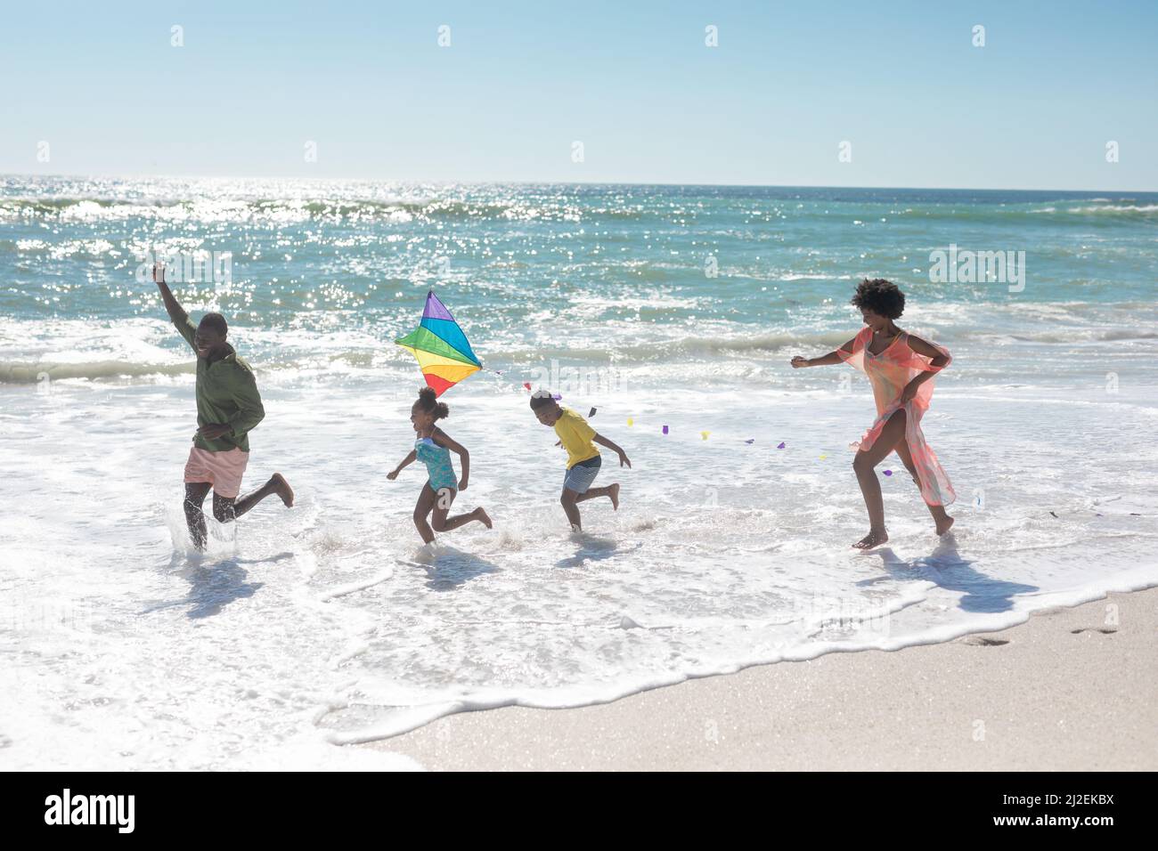 Felice famiglia afroamericana che corre con aquilone sulla spiaggia durante la giornata di sole Foto Stock