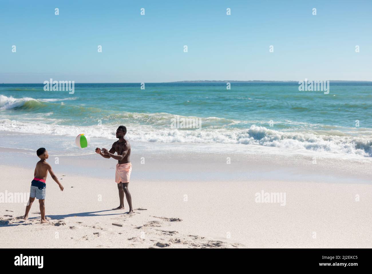 Tutta la lunghezza di padre e figlio afro-americani che giocano con la palla in spiaggia il giorno di sole Foto Stock