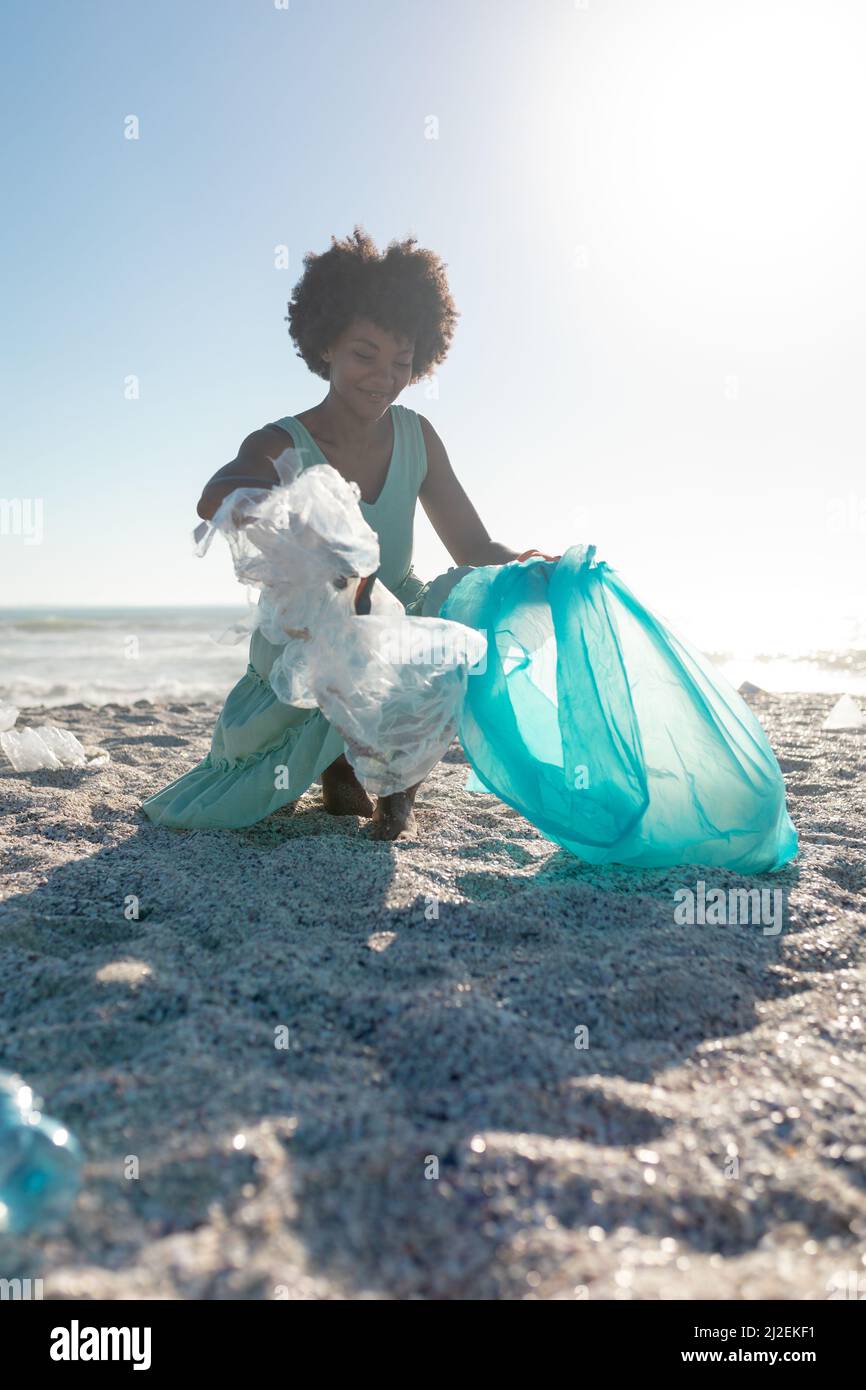 Donna afroamericana che raccoglie rifiuti e raccogliendo in sacchetto di plastica alla spiaggia contro cielo limpido Foto Stock