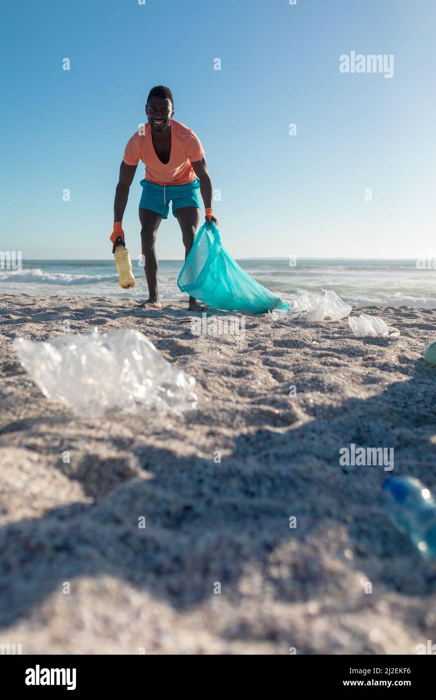 Felice uomo afroamericano raccolta rifiuti in sacchetto di plastica in spiaggia contro cielo blu chiaro Foto Stock