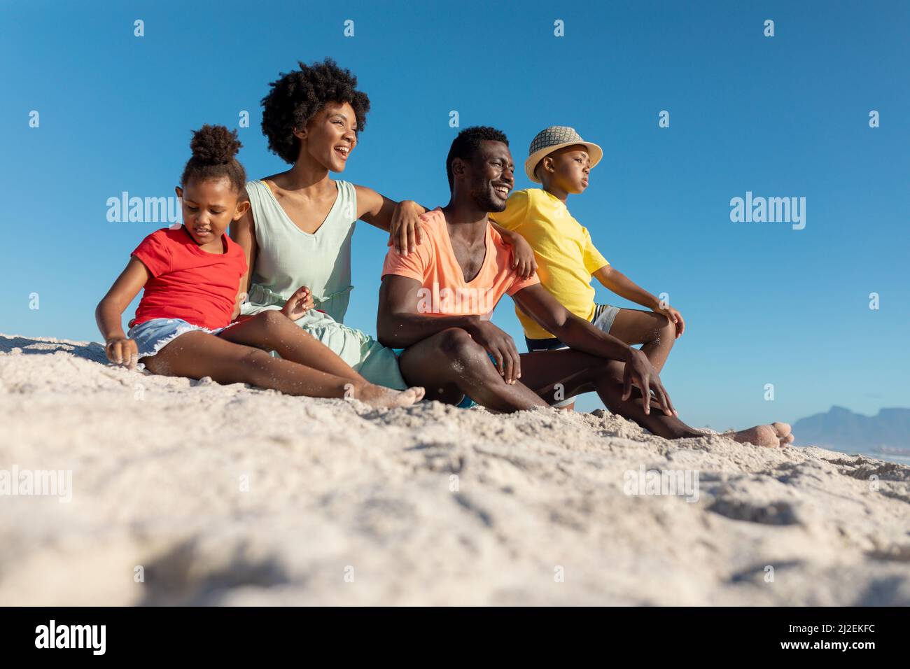 Vista a livello della superficie della famiglia afro-americana seduta sulla sabbia sulla spiaggia contro il cielo blu chiaro Foto Stock