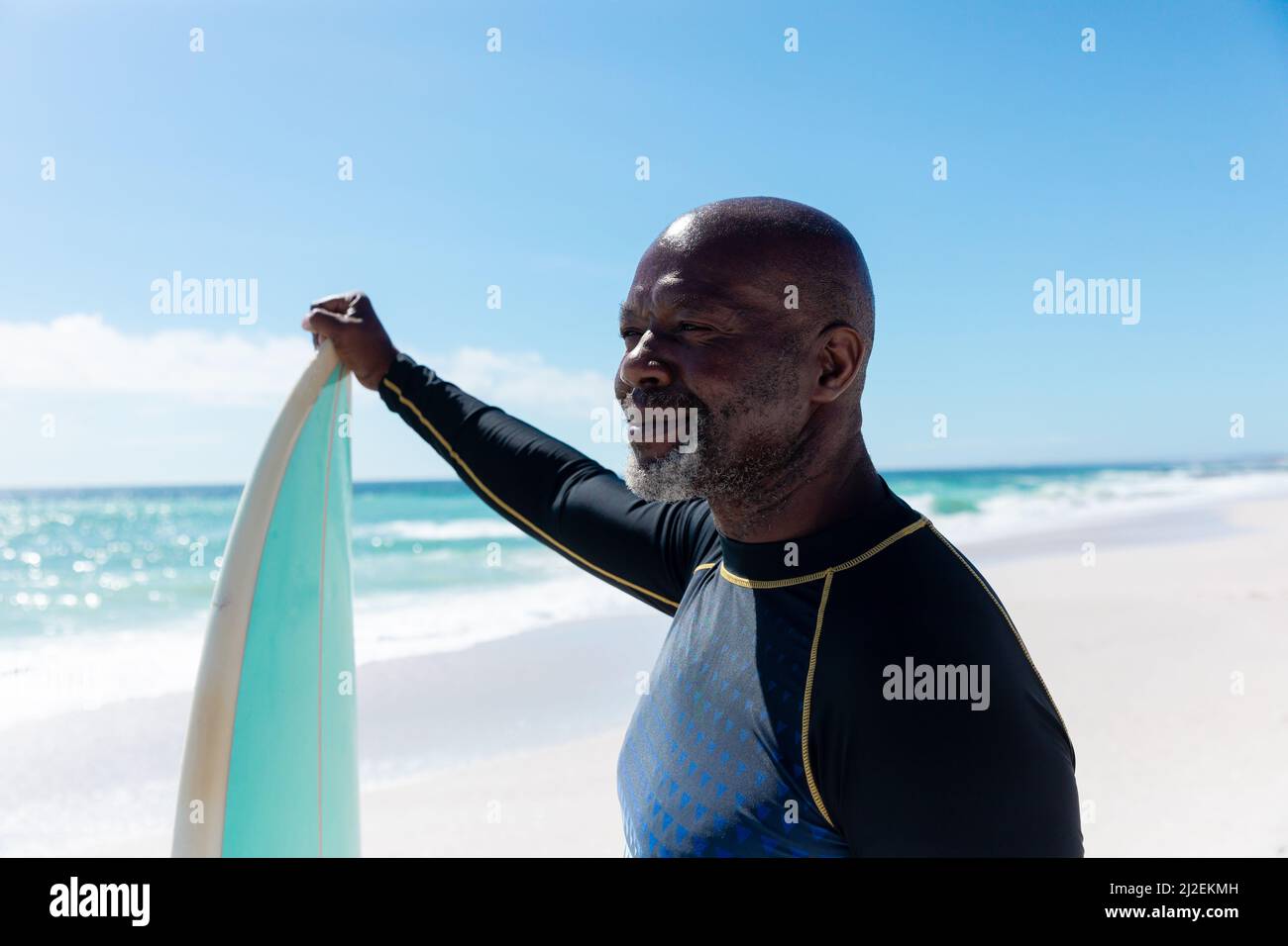 Sorridente uomo anziano afro-americano che tiene la tavola da surf guardando via alla spiaggia in giorno di sole Foto Stock