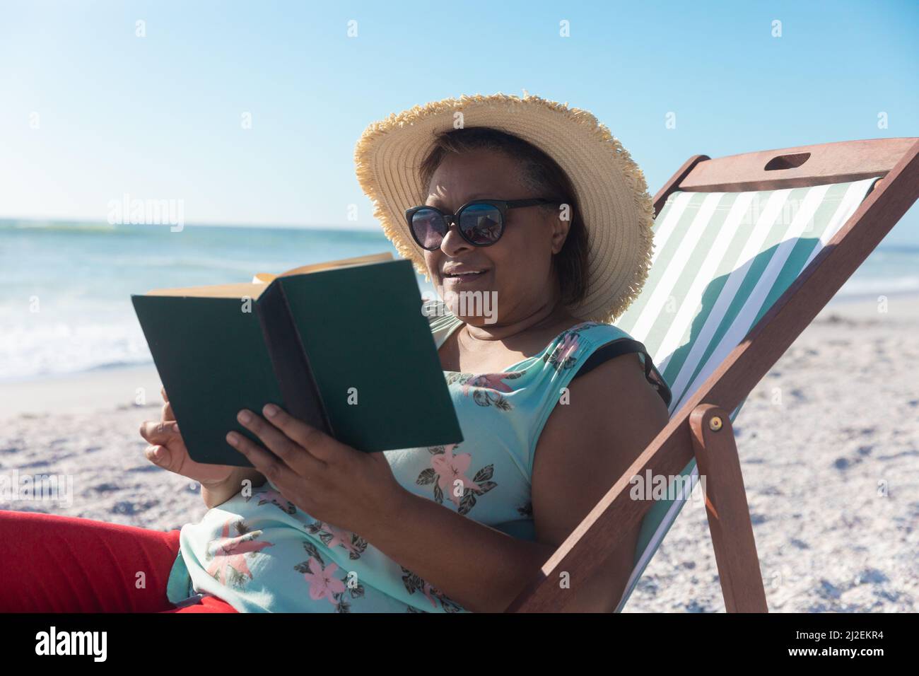 African american pensionato Senior donna lettura libro mentre si siede su sedia pieghevole in spiaggia Foto Stock