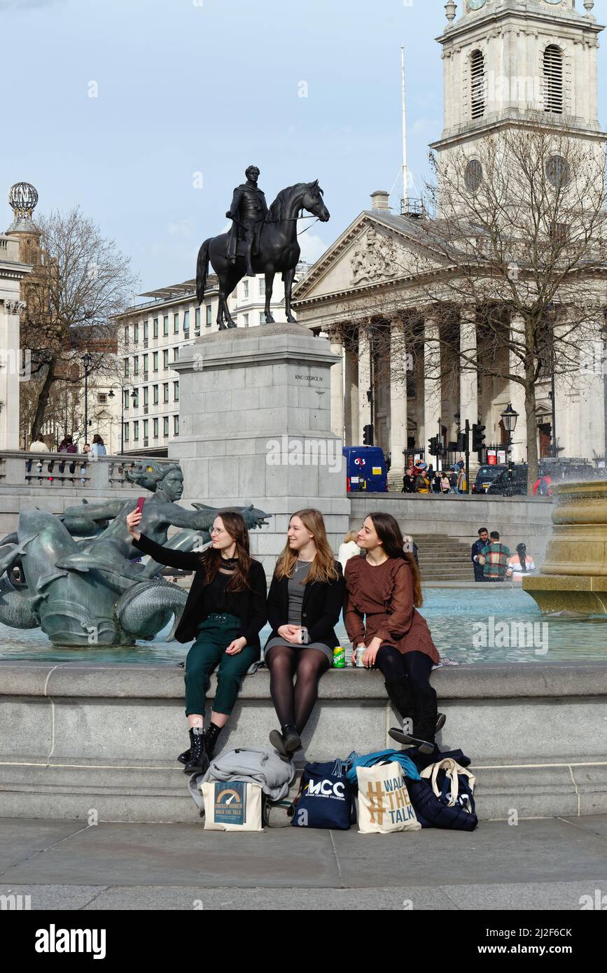 Tre ragazze attraenti giovani seduti accanto alla fontana in Trafalgar Square prendendo un selfie con una grande statua equestre sullo sfondo Londra UK Foto Stock