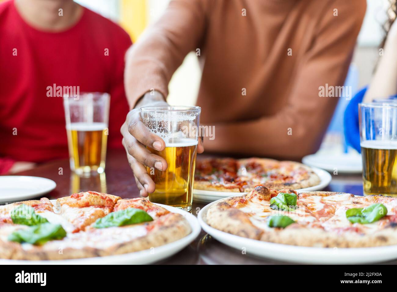 Primo piano del ragazzo africano che prende a mano un bicchiere di birra al ristorante pizza - amici multietnici che si divertono a mangiare pizza e bere birra al pub Foto Stock