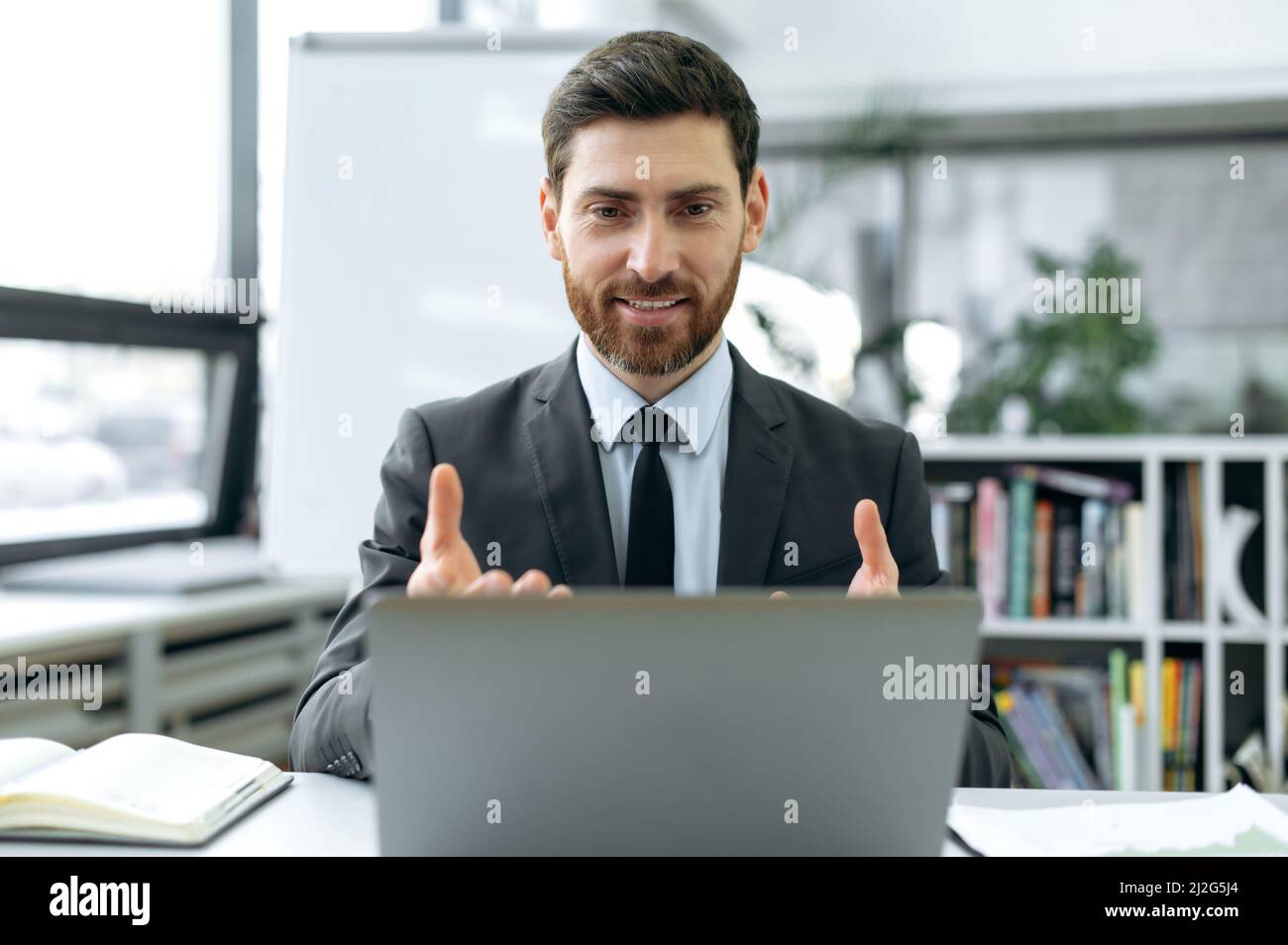 Concentrato uomo d'affari caucasico, ingegnere della sicurezza delle informazioni, responsabile assicurativo, sedersi in ufficio moderno, avere video incontro con il collega utilizzando il laptop, spiegare la strategia di lavoro, gesti mani Foto Stock