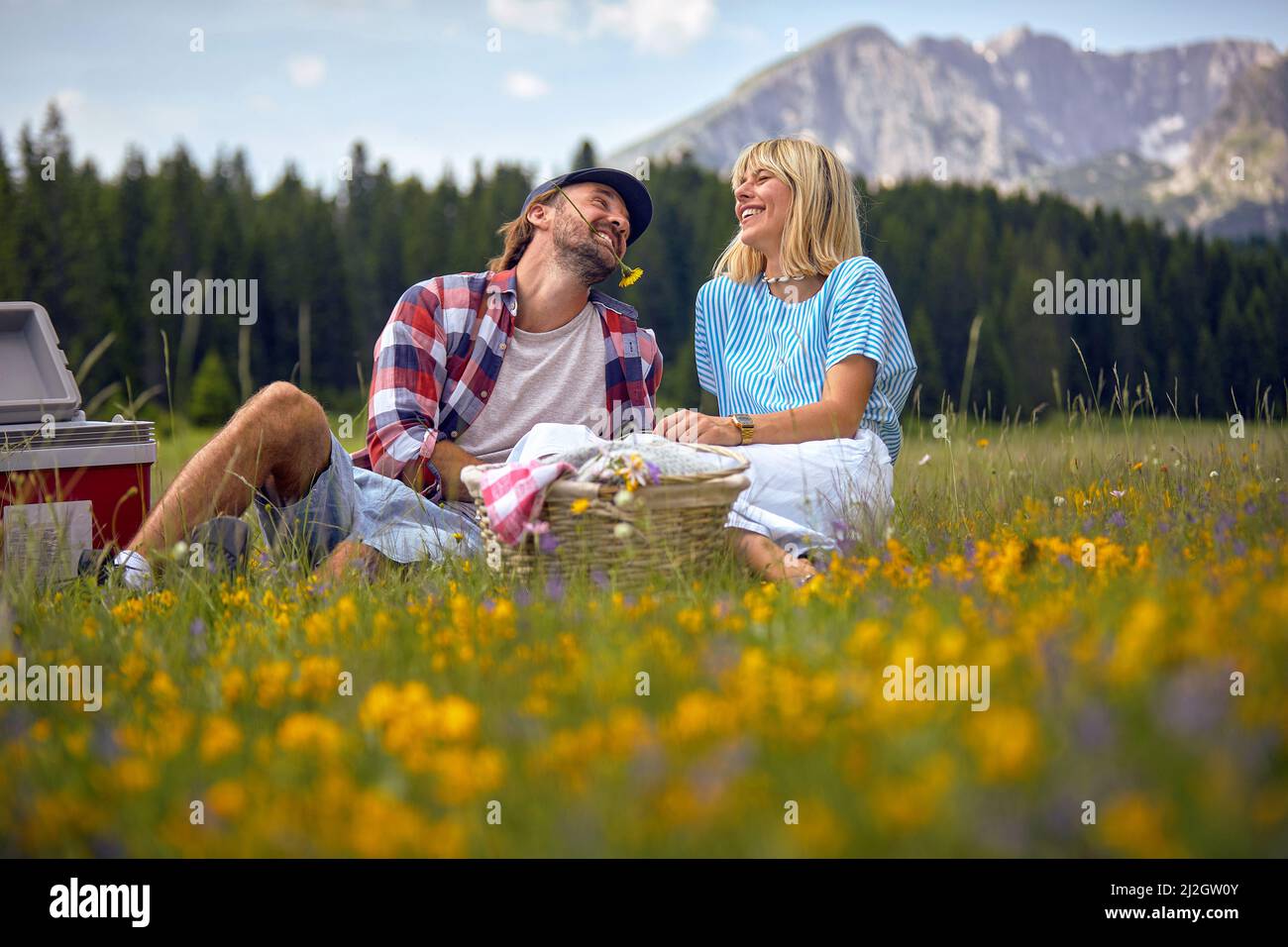 Giovane coppia che si diverte sul prato, picnic tempo. Uomo con fiore in bocca. Divertimento, convivenza, stile di vita, concetto di natura. Foto Stock