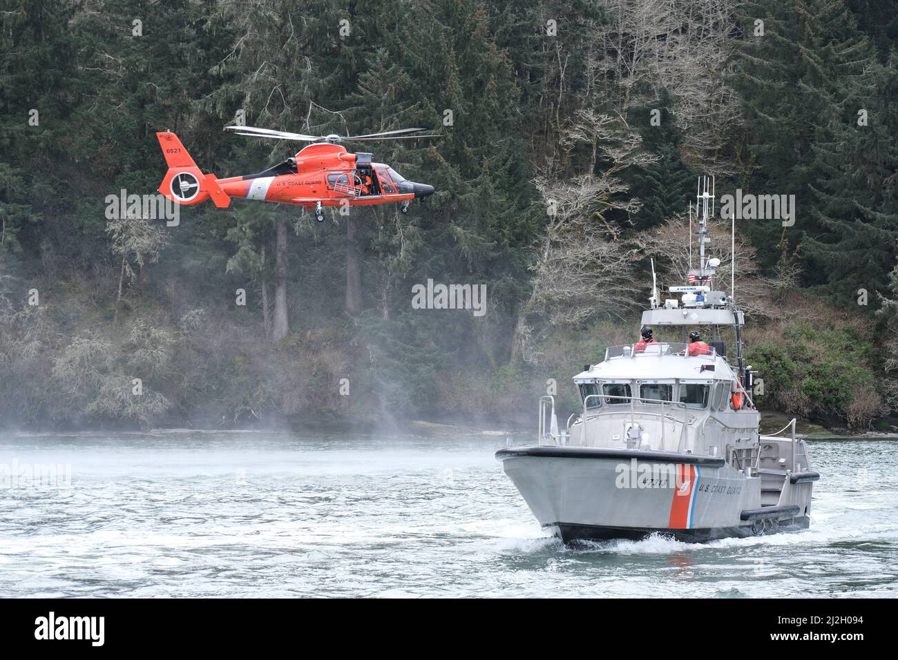 L'equipaggio a bordo di un motonavalvascelli di 47 piedi dalla stazione della Guardia Costiera del fiume Umpqua conduce l'addestramento del paranco con un equipaggio MH-65 Delfino Helicopter dall'Air Station North Bend nel fiume Umpqua vicino a Winchester Bay, Oregon, 15 marzo 2022. Le operazioni di addestramento sono tenute regolarmente per assicurare che le capacità di ricerca e di salvataggio degli equipaggi della Guardia Costiera rimani all'alto livello previsto da loro. Foto Stock