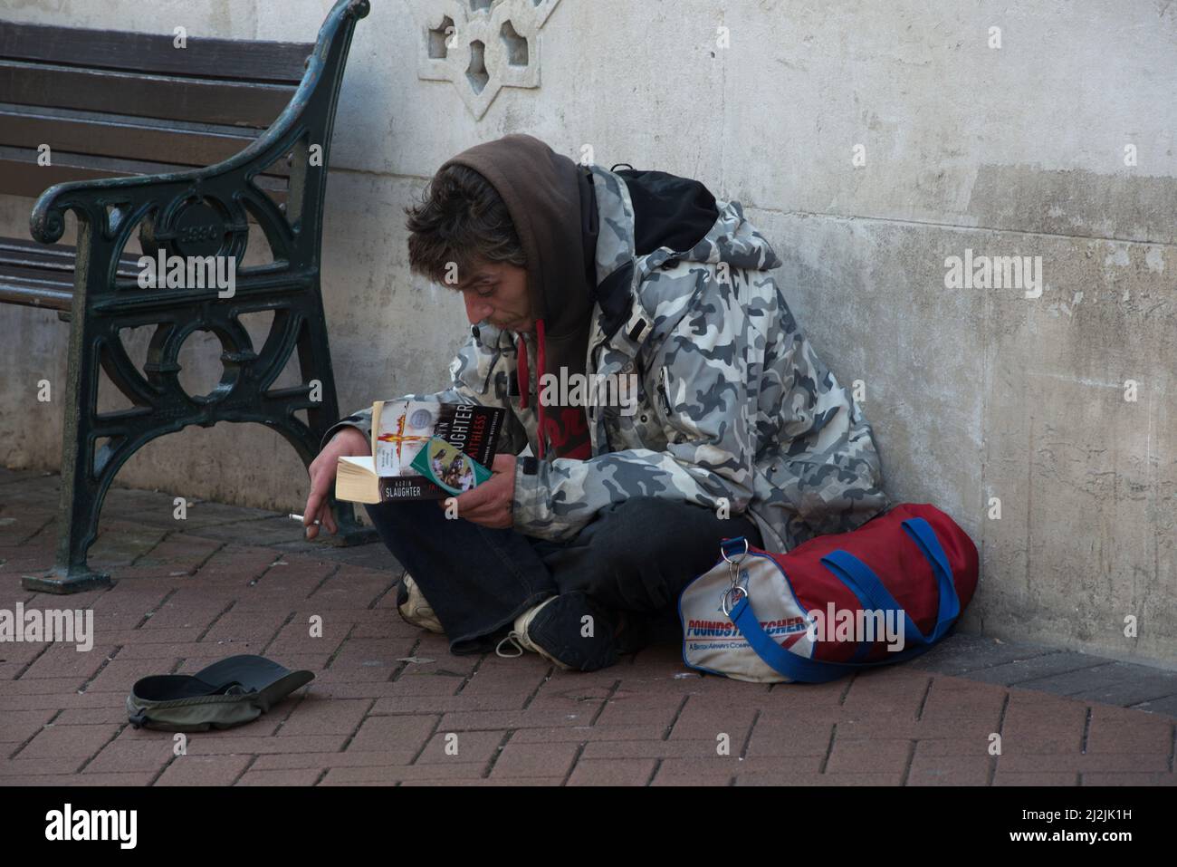 L'uomo senza tetto legge un libro mentre fuma una sigaretta per le strade di Bournmouth Foto Stock