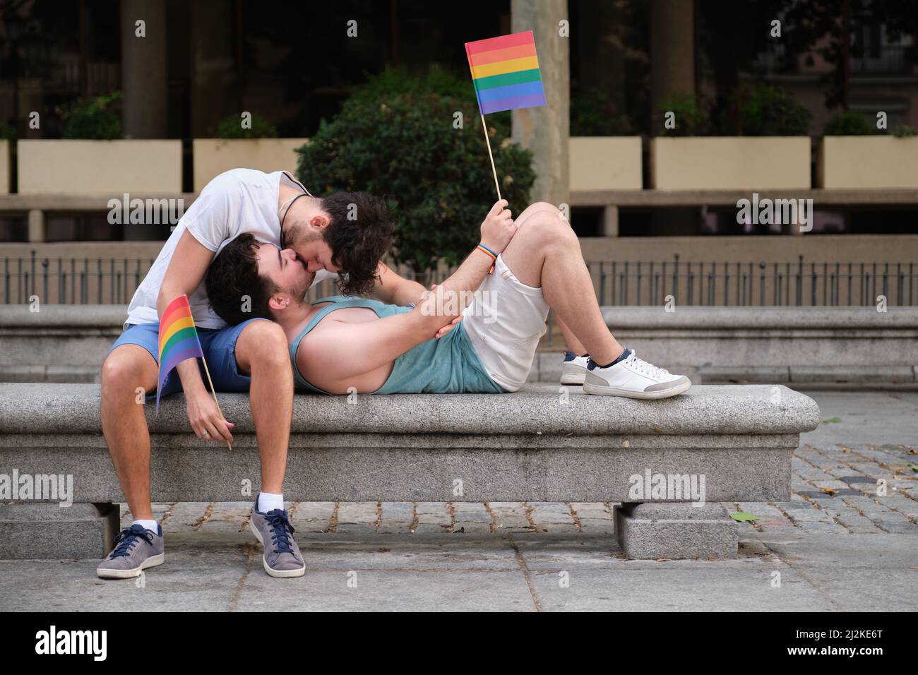 Coppia gay ridendo e abbracciando seduti su una panchina che regge bandiere LGBT. Foto Stock