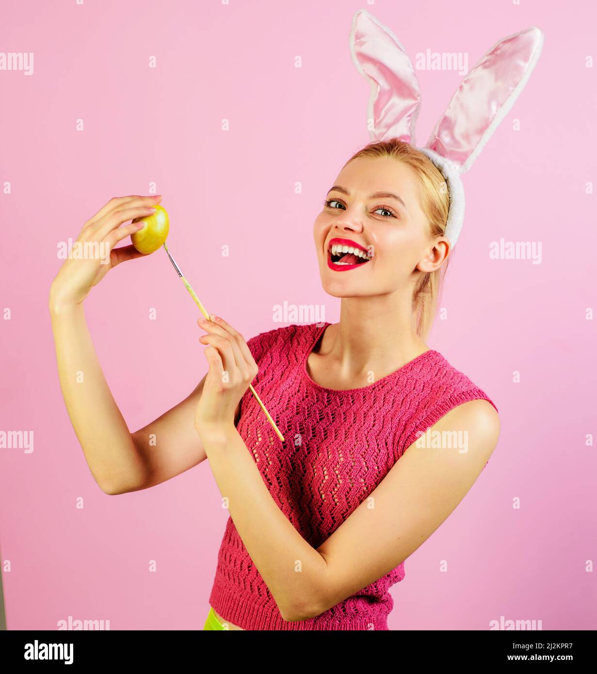 Pasqua ragazza in coniglietto orecchie pittura uova. Coniglio donna con uovo colorato e pennello. Foto Stock