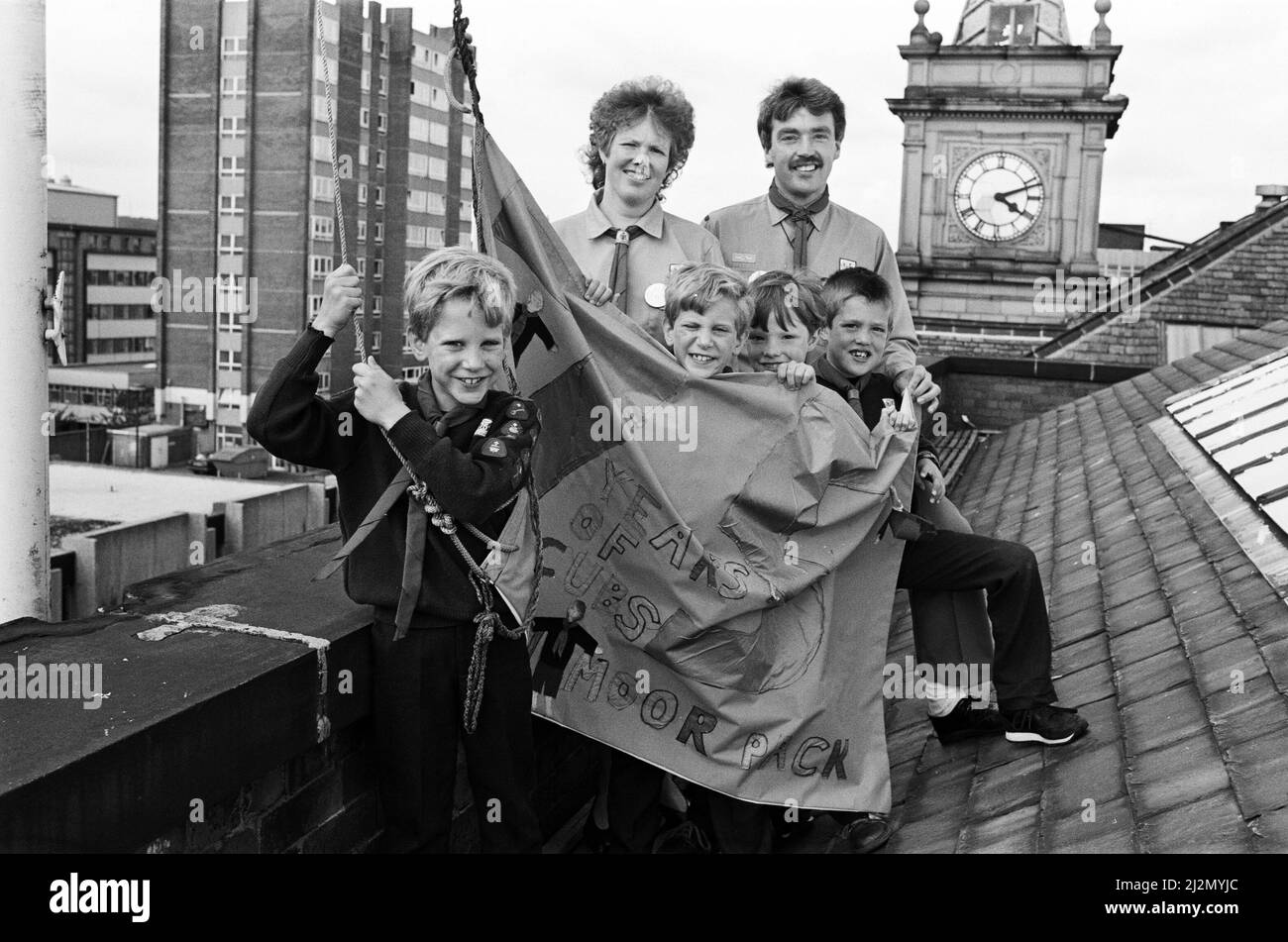 Volando la bandiera... questo è ciò che sta facendo il pack Netherton Moor Cub per festeggiare il 75th compleanno del movimento - e visto con la bandiera del gruppo sono (davanti, da sinistra) nove anni gemelli Edward e James Broadbent e fratelli Edward, 10 e Gareth Johns, otto. Nella foto sono raffigurati anche i genitori e i capi Cub Susan e Kevin Johns. Ai cubani di tutto il paese è stato chiesto di progettare la propria bandiera e poi trovare luoghi insoliti per volare loro. La bandiera Netherton viene volata dall'edificio Co-op di New Street, una delle più alte del centro della città di Huddersfield. 24th giugno 1991. Foto Stock