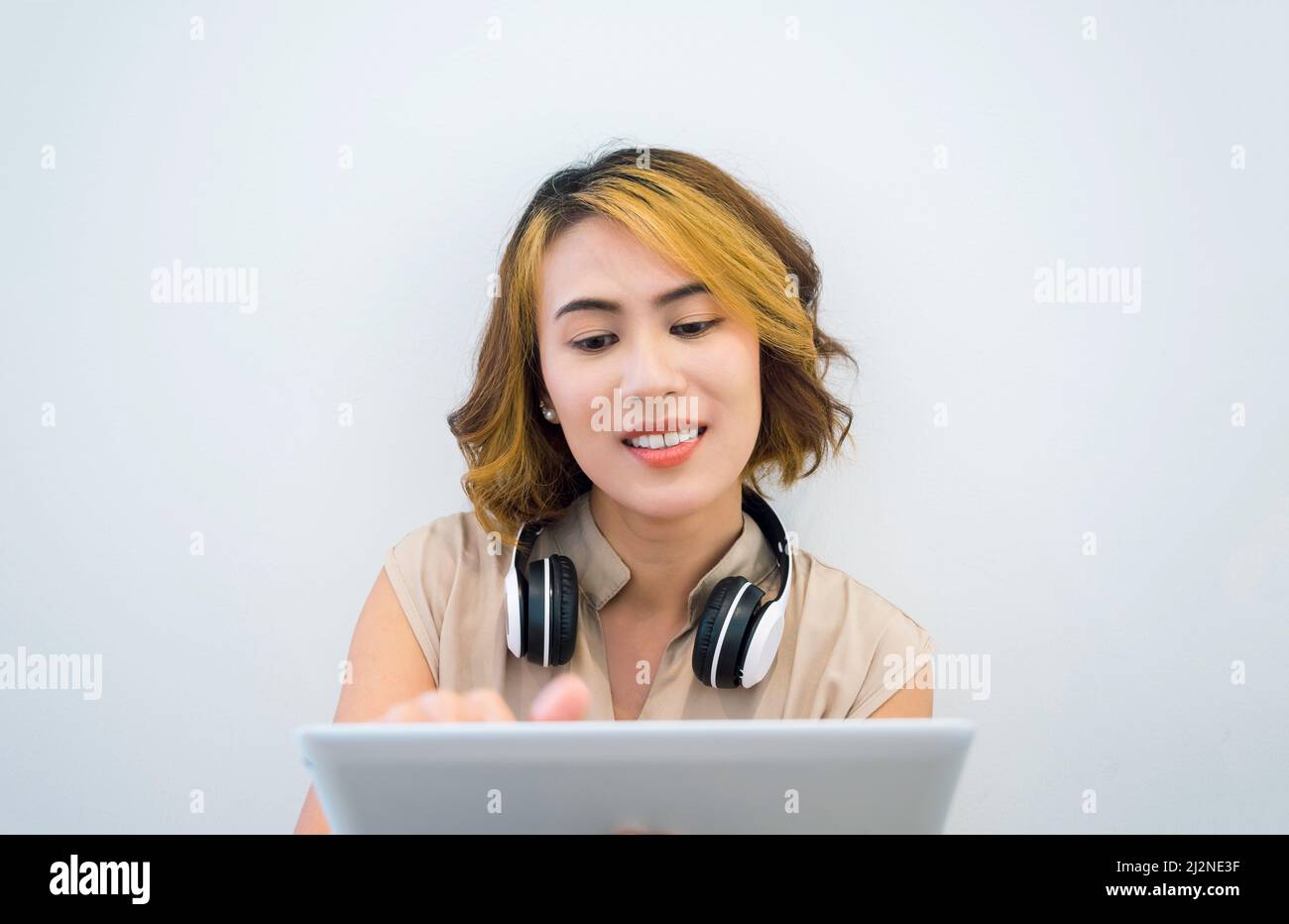 Donna asiatica felice capelli corti in camicia senza maniche beige guardando il tablet e toccando sullo schermo con cuffie wireless bianche sul collo su parete bianca ba Foto Stock