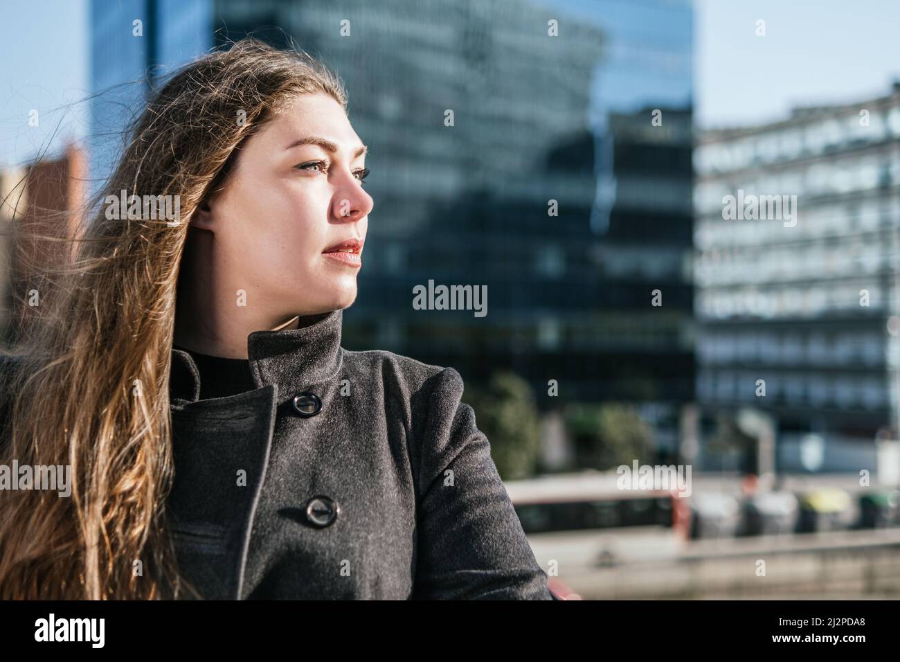 Donna seria con lunghi capelli volanti indossando un caldo cappotto in piedi contro la torre di vetro sfocato e ammirando le strade della città Foto Stock