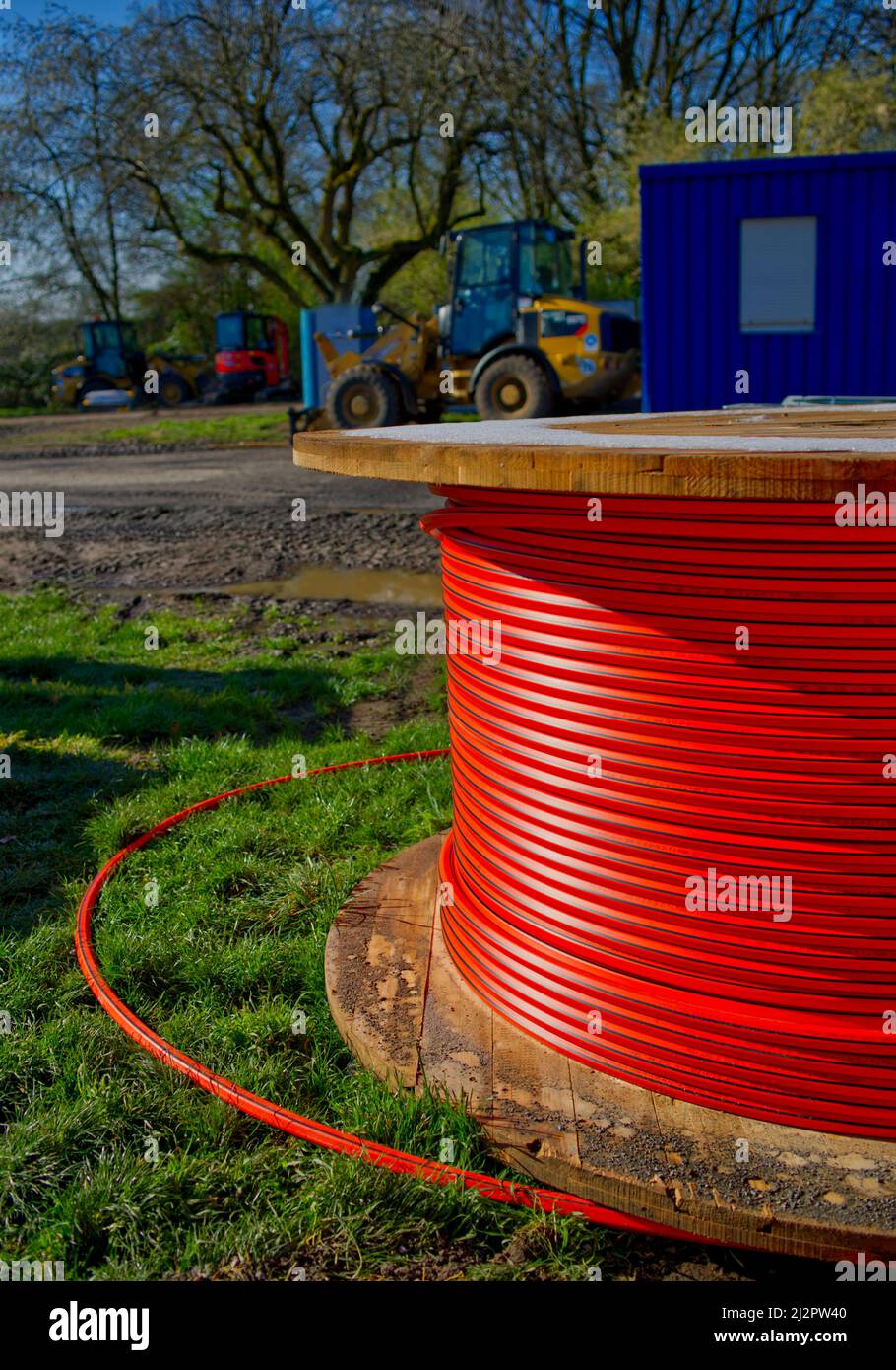 Enorme avvolgicavo di cavo in fibra ottica rossa in un cantiere con veicoli in cantiere e un blu sullo sfondo Foto Stock Enorme avvolgicavo di cavo in fibra ottica rossa in un cantiere con veicoli in cantiere e un blu sullo sfondo Foto Stock