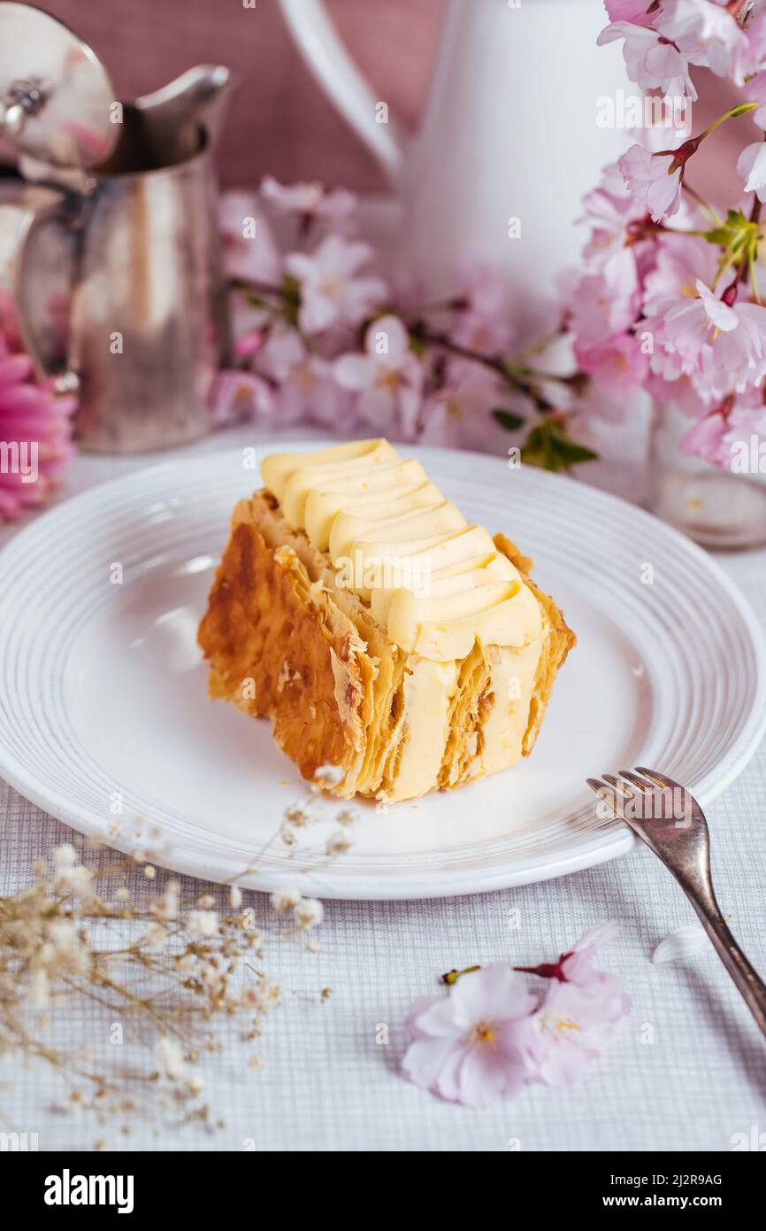 Dessert francese chiamato Mille Feuille o torta Napoleone decorato con fiori in fiore su sfondo luminoso. Torta gustosa a strati con crema alla vaniglia e pu Foto Stock