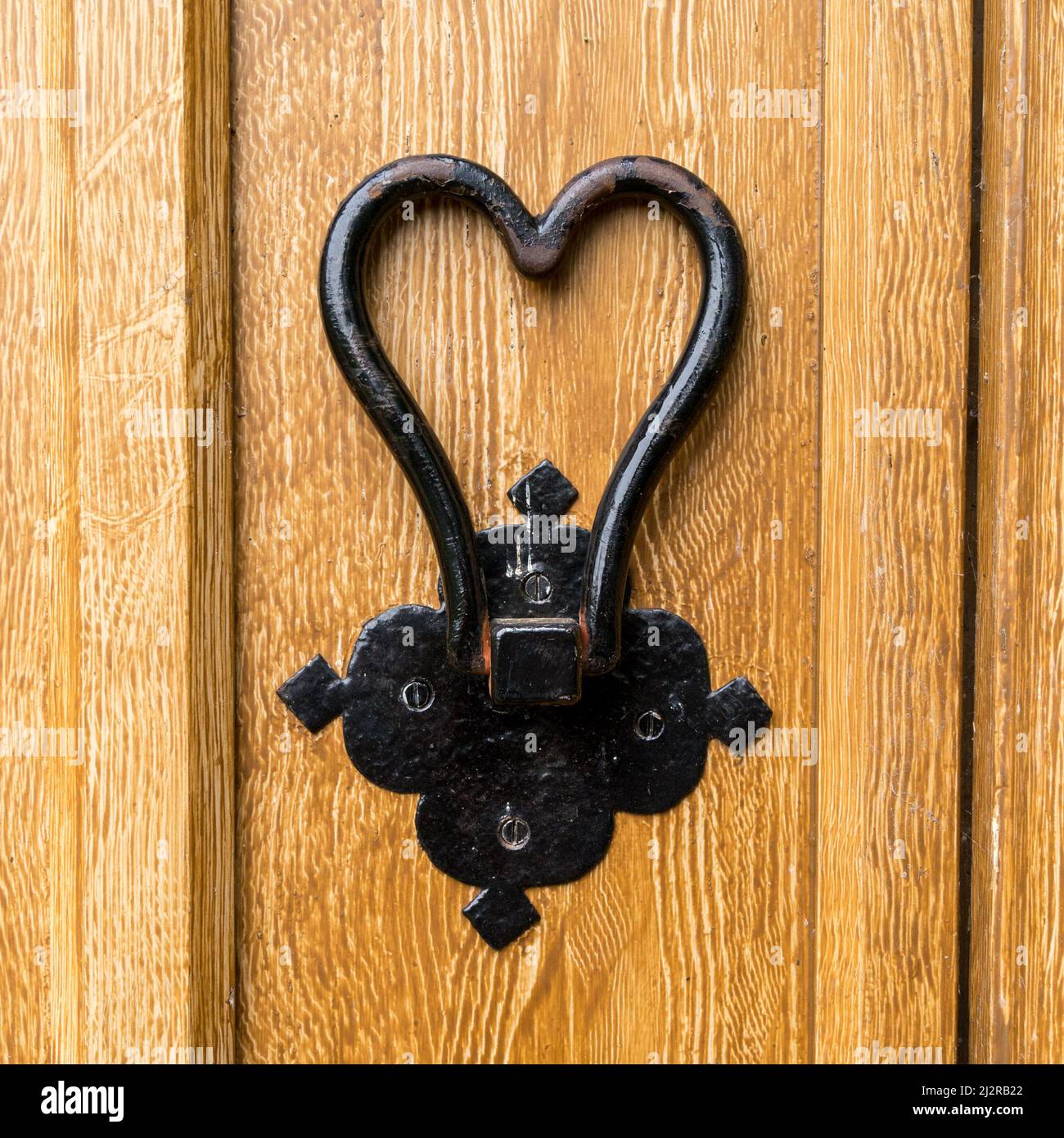 Battente di porta a forma di cuore in ferro battuto dipinto di nero su porta in legno verniciato con effetto di finta grana di legno marrone chiaro. Foto Stock