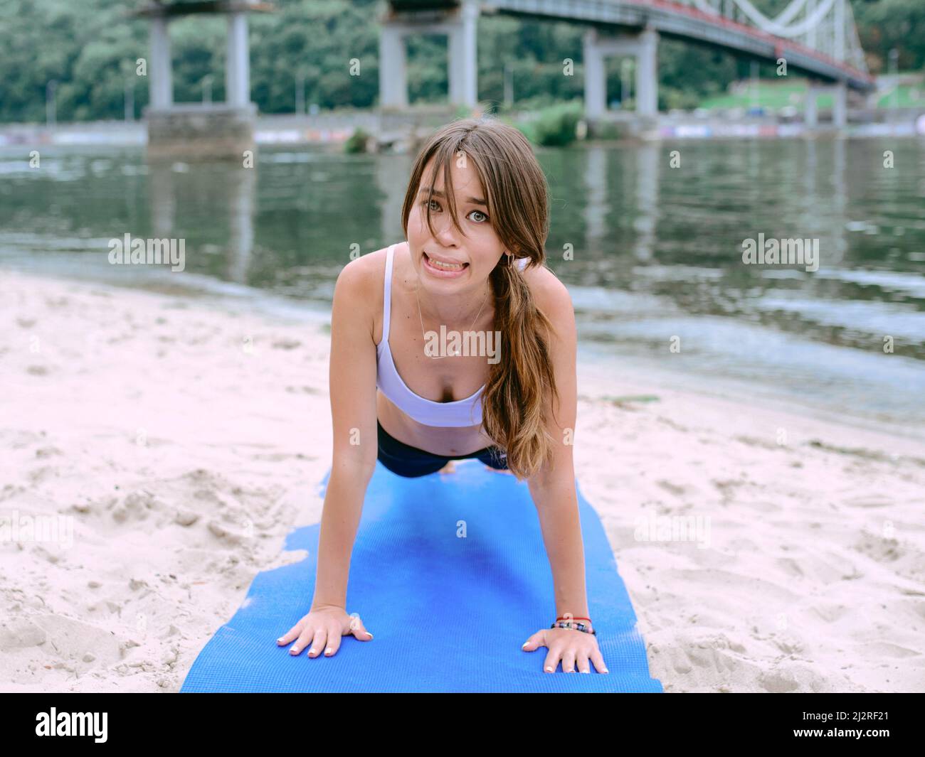 giovane donna in abbigliamento sportivo che inizia lo yoga e ha problemi con lo yoga asanas all'aperto sulla spiaggia vicino al fiume. Yoga e concetto di sport Foto Stock