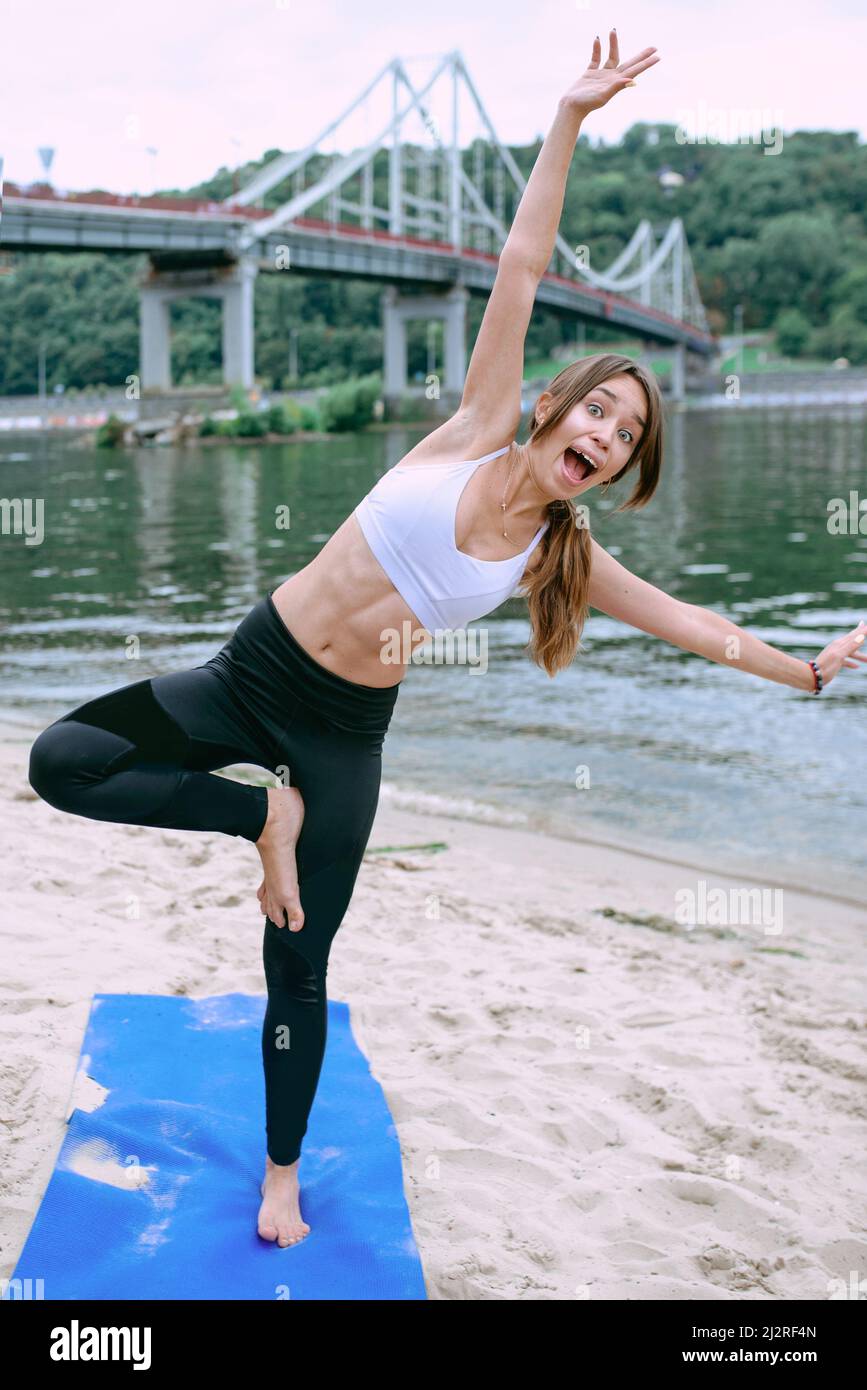 giovane donna in abbigliamento sportivo che inizia lo yoga e ha problemi con lo yoga asanas all'aperto sulla spiaggia vicino al fiume. Yoga e concetto di sport Foto Stock
