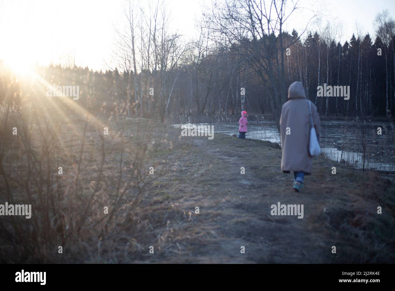 Una ragazza cammina al tramonto con un bambino. Passeggiata sul lago nella foresta. Giovane madre in una giacca calda. Riprese dal retro. Foto Stock