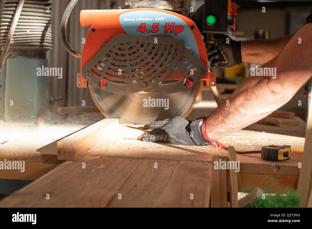 le mani di un giovane falegname che lavora su una sega circolare che taglia un asse di legno in un'officina di carpenteria usando guanti di sicurezza neri Foto Stock