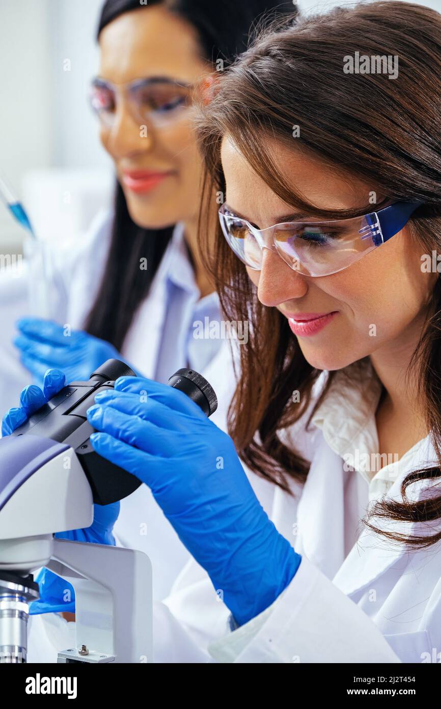 Giovane scienziata femminile al microscopio mentre il suo collega lavora nel settore sanitario di base Foto Stock