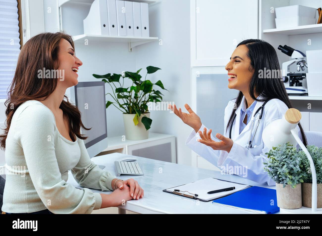 Ride femmina medico e paziente cliente signora discutere qualcosa mentre si siede al tavolo nel centro medico settore sanitario Foto Stock