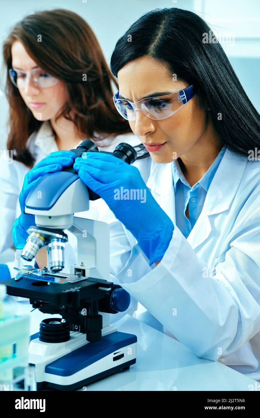 Giovane scienziata femminile che usa il microscopio mentre la sua collega che lavora nell'industria farmaceutica di base Foto Stock