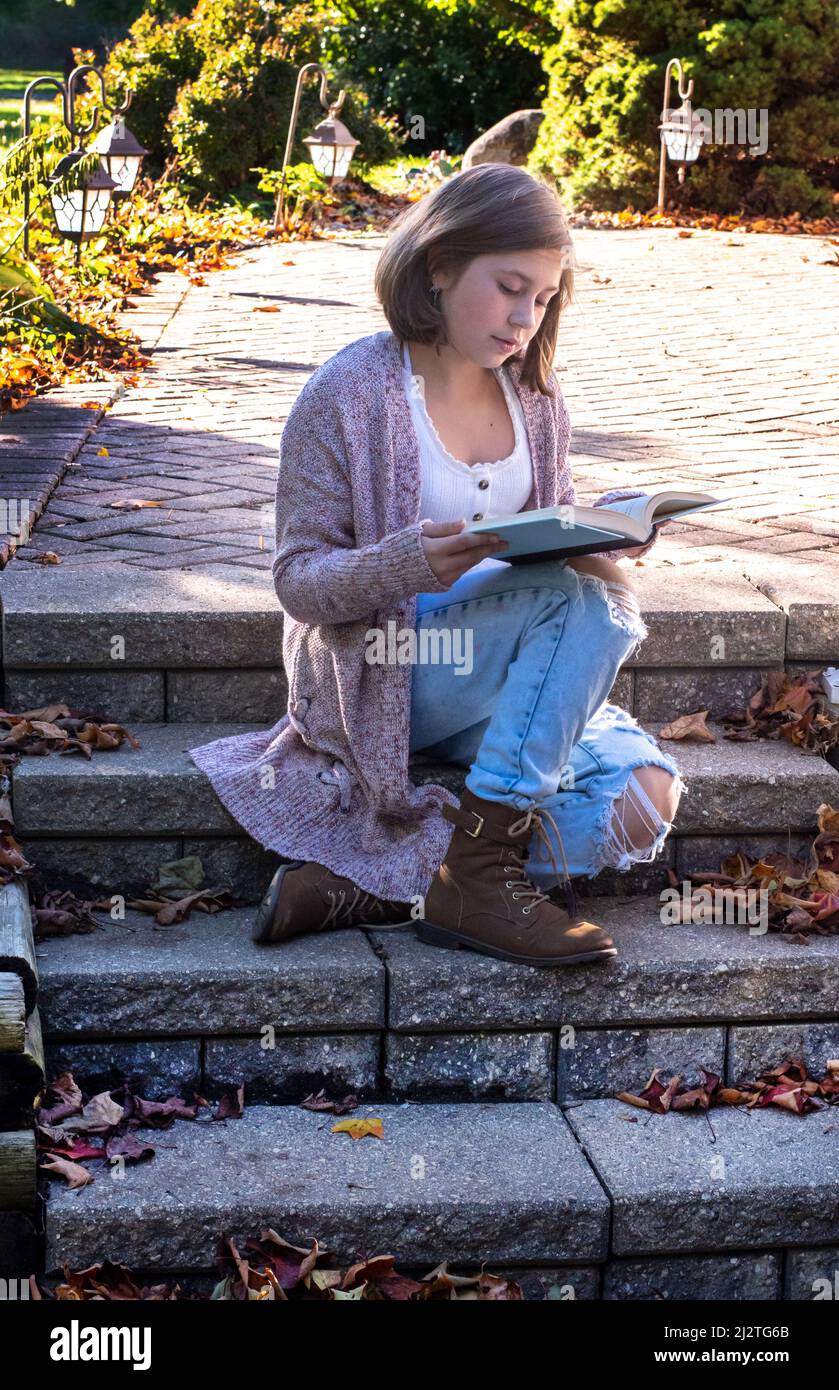 La ragazza giovane siede sui suoi passi del portico che leggono un libro su un giorno di caduta grazioso Foto Stock