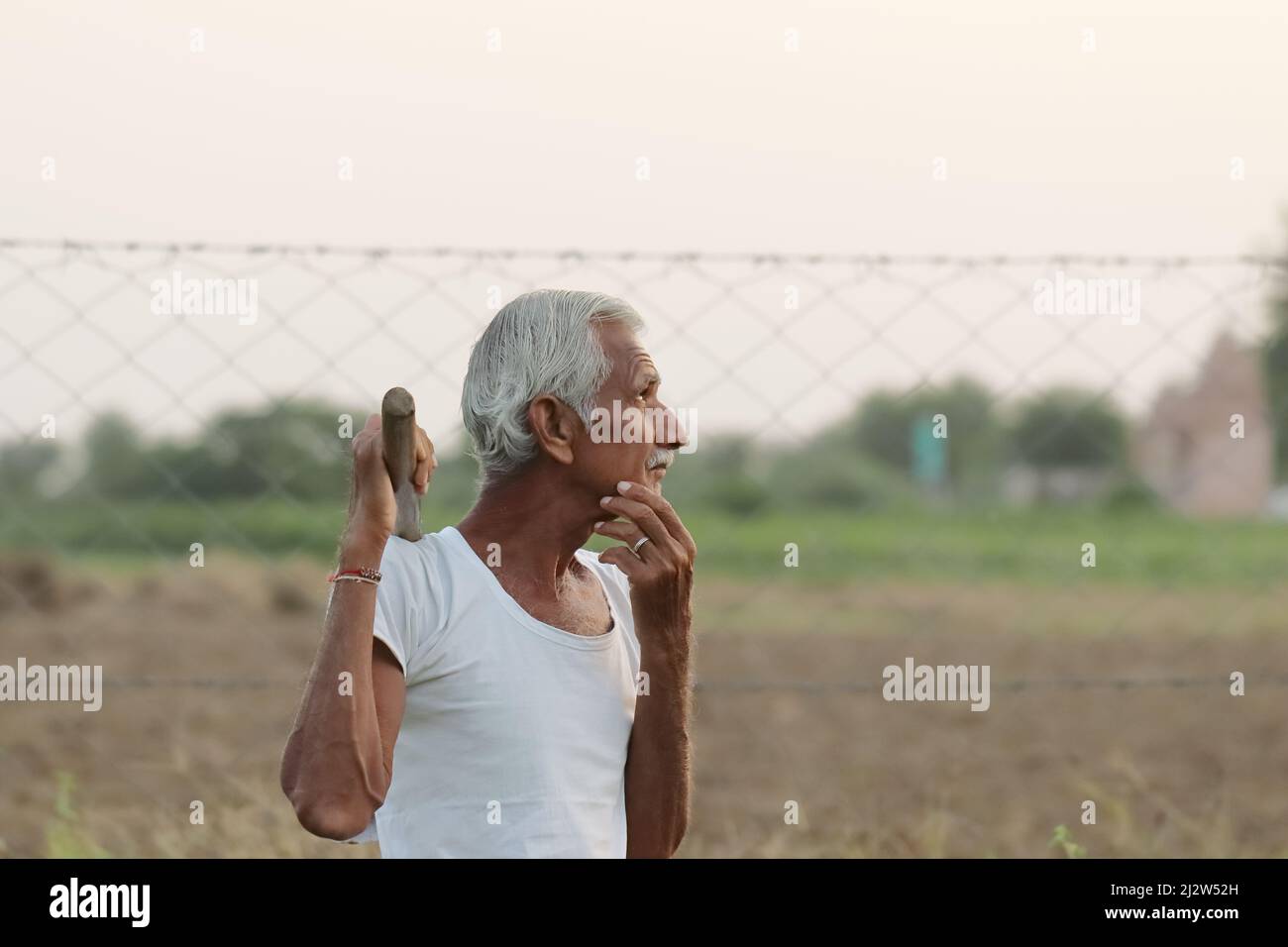 Primo piano di una foto di un agricoltore indiano anziano che pensa a qualcosa mentre tiene una pala in mano al tramonto Foto Stock