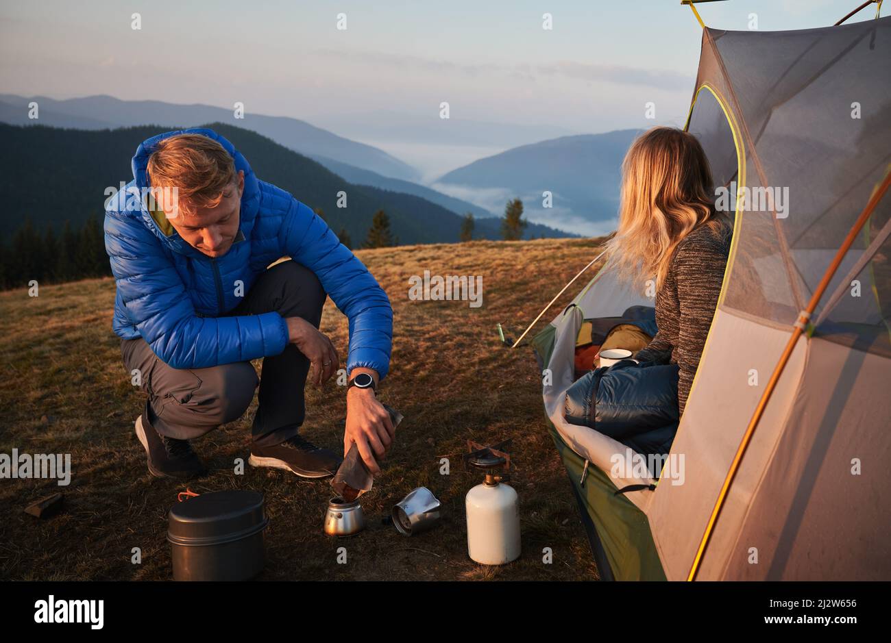 Mattina in estate tenda accampamento all'aria fresca. Uomo che prepara la colazione per la sua ragazza. Nebbia mattutina sulle colline di montagna sullo sfondo. Concetto di escursioni, campeggio e relazioni. Foto Stock
