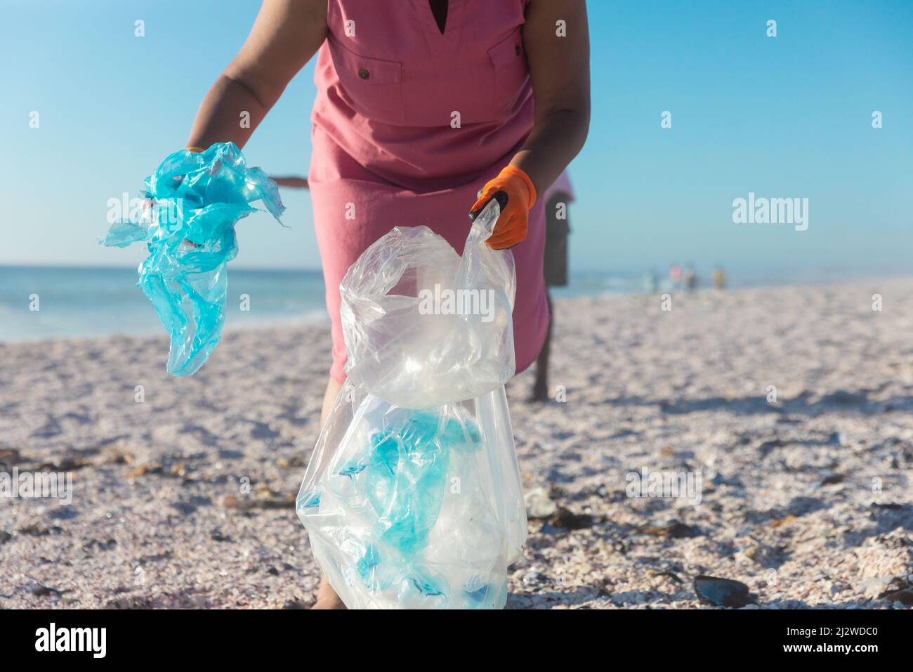 Donna anziana in pensione afro-americana che raccoglie rifiuti di plastica in borsa alla spiaggia il giorno di sole Foto Stock