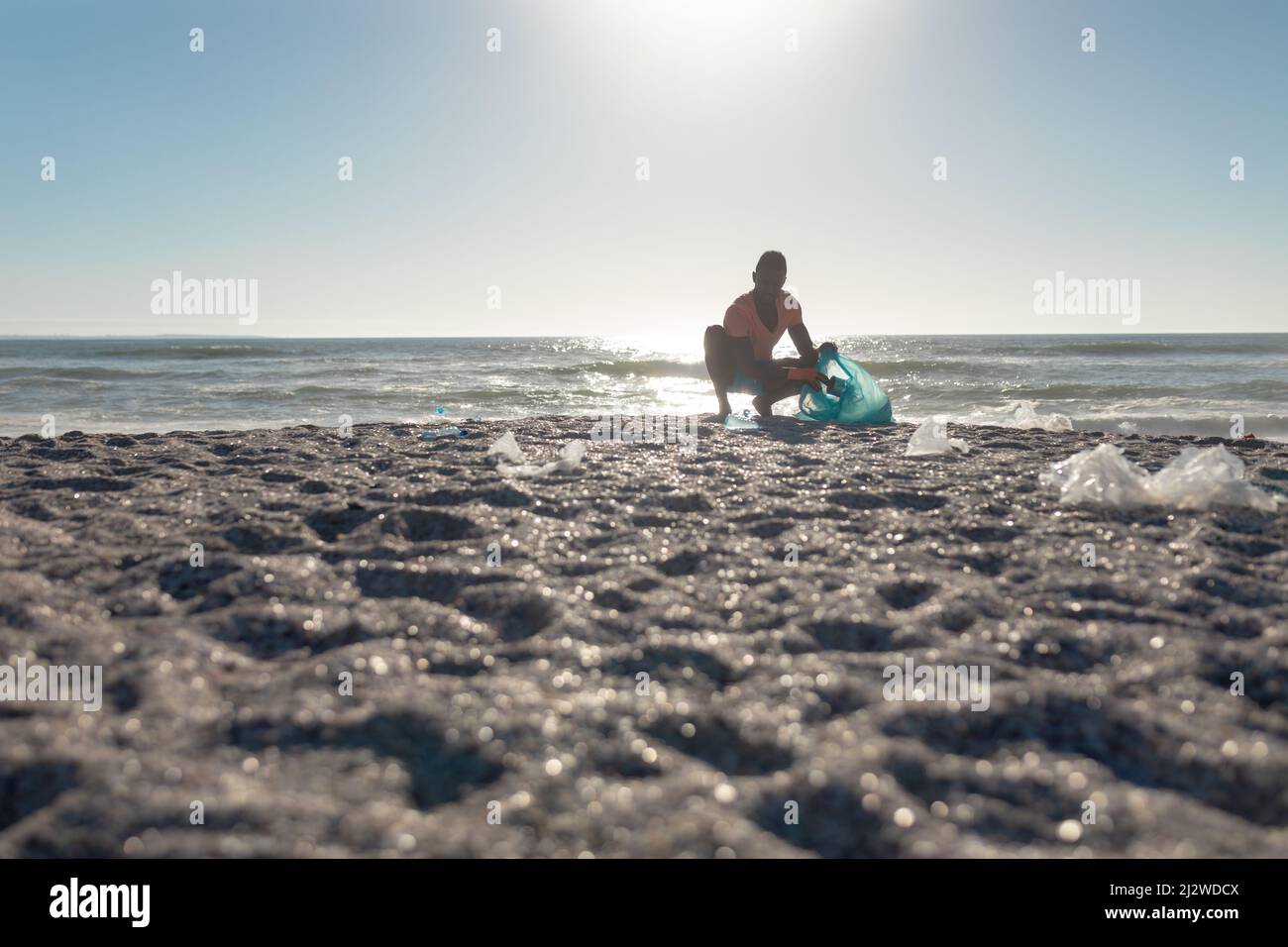 Vista a livello della superficie dell'uomo afro-americano accovacciato mentre raccoglie i rifiuti in borsa alla spiaggia Foto Stock