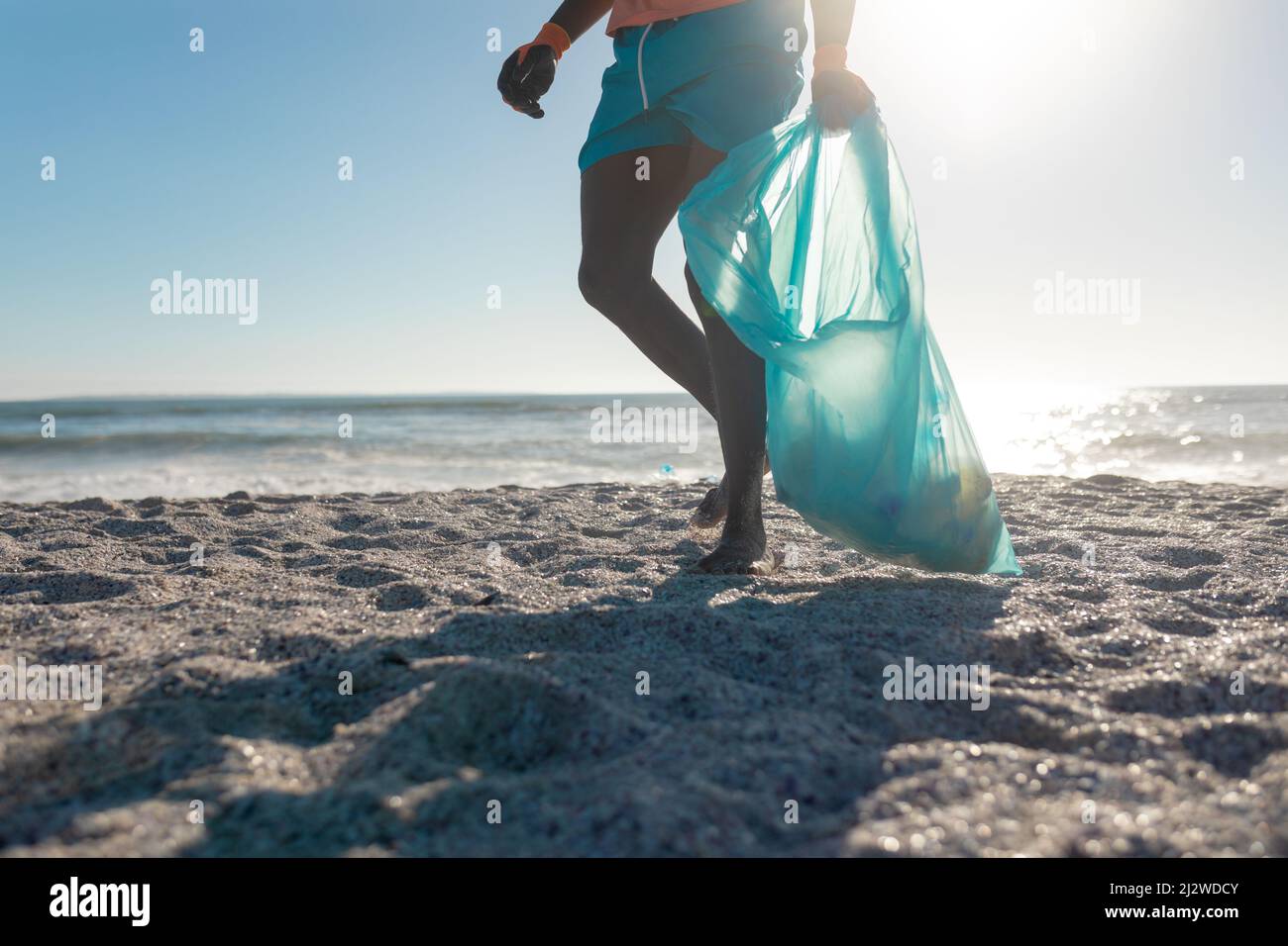 Parte bassa di uomo che trasporta la borsa di plastica con i rifiuti che camminano in spiaggia in giorno di sole Foto Stock