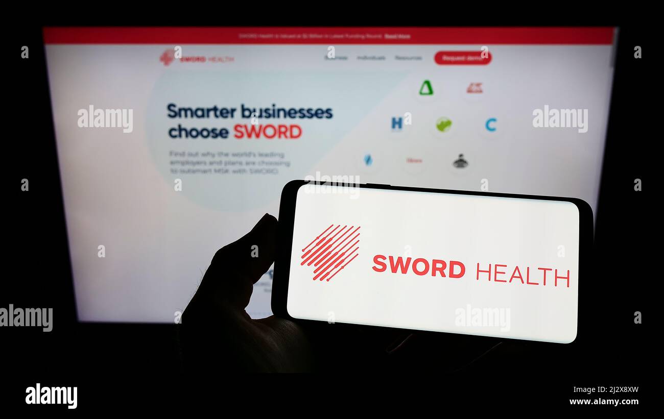 Persona che tiene il telefono cellulare con il logo della società di e-Health SWORD Health Inc. Sullo schermo di fronte alla pagina web aziendale. Mettere a fuoco sul display del telefono. Foto Stock