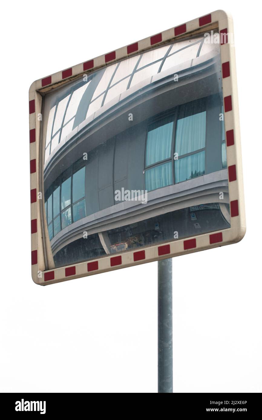Uno specchio stradale riflette un'immagine distorta della facciata di un edificio grigio con finestre. La cornice dello specchio stradale è bianca e rossa. Il piede è Foto Stock