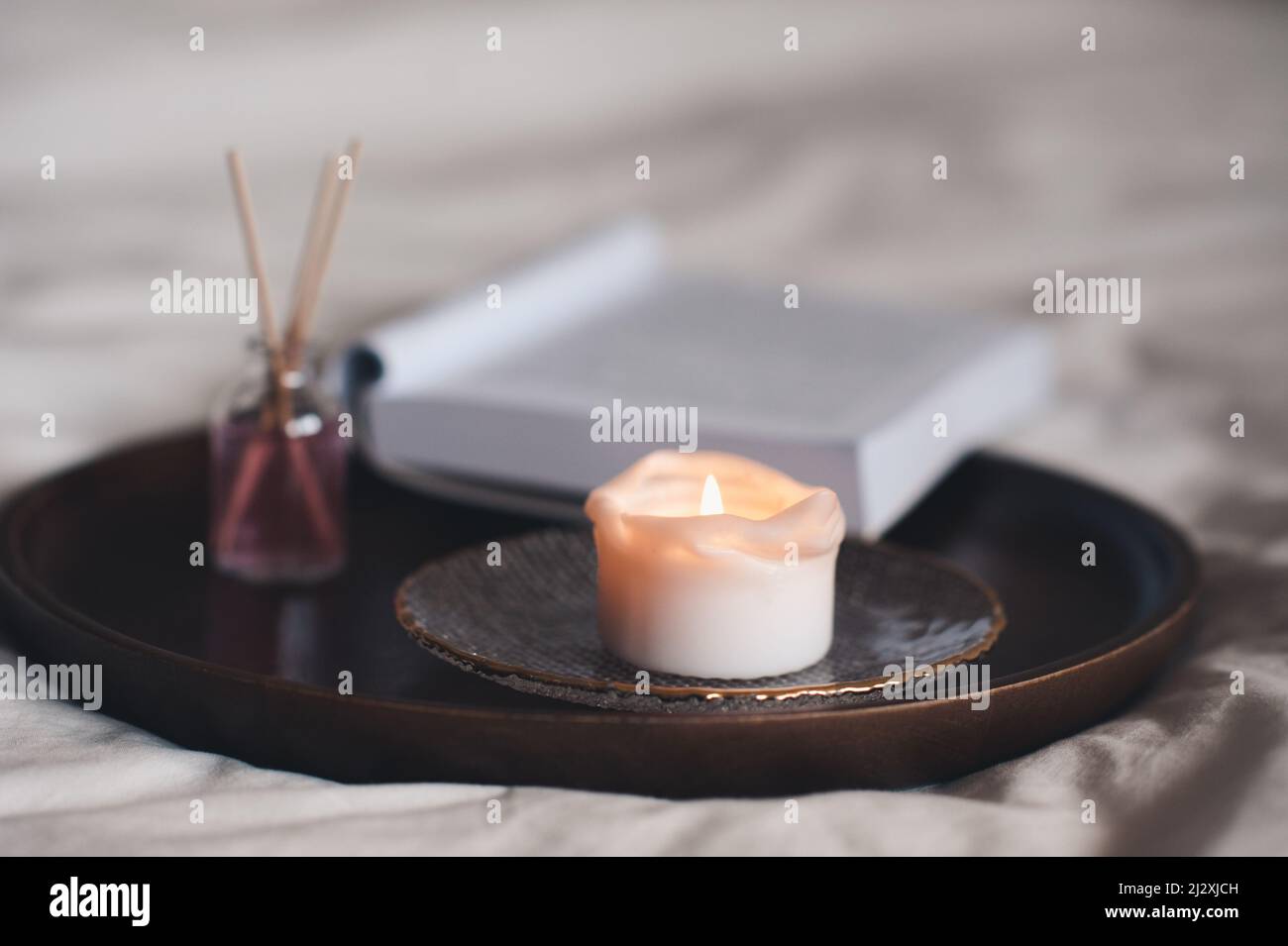 Home fragranza liquida in bottiglia di vetro con bastoni di legno e candela profumata con libro di carta aperto su vassoio in letto da vicino. Foto Stock