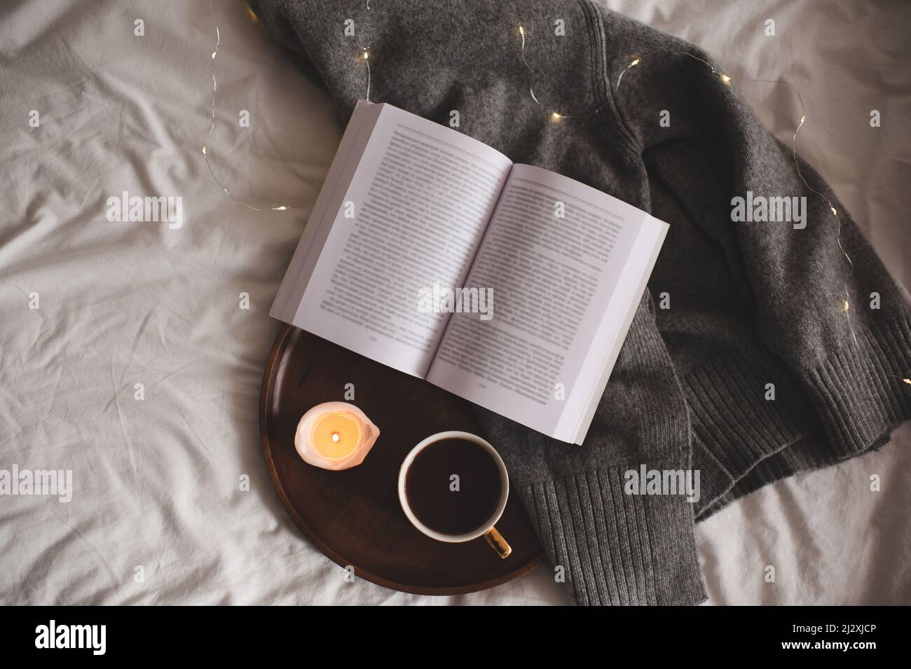 Tazza di caffè con stampa leopardo che rimane su vassoio di legno con profumata candela bruciante e libro di carta aperto su tessuto lavorato a maglia a letto a casa. Buon mattino Foto Stock
