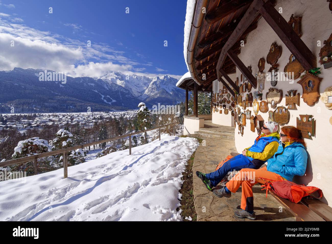 L'uomo e la donna mentre camminano si siedono di fronte alla chiesa commemorativa della guerra con le montagne di Wetterstein sullo sfondo, Kramerplateauweg, Garmisch, Alpi di Ammergau, Werdenfelser Land, Baviera superiore, Baviera, Germania Foto Stock