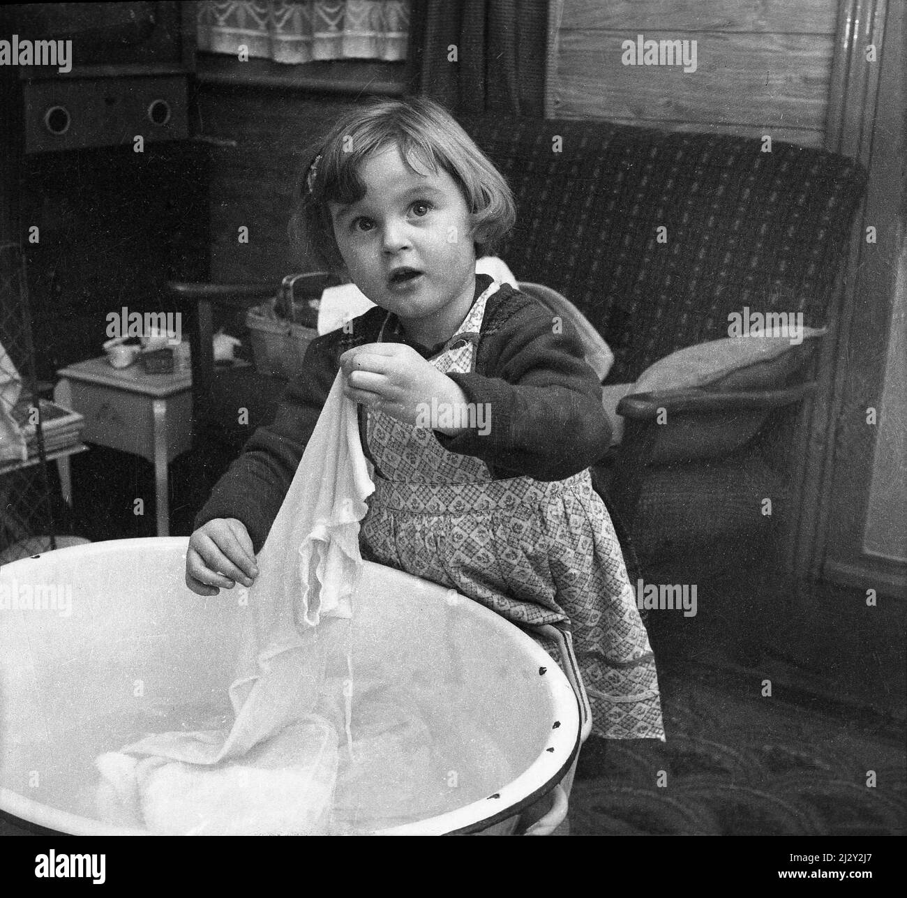 1961, hisotrical, in piedi in una stanza anteriore, bambina che tiene su un capo di vestiario che è stato lavato in un bacino o ciotola di smalto, Stockport, Manchester, Inghilterra, Regno Unito. Foto Stock