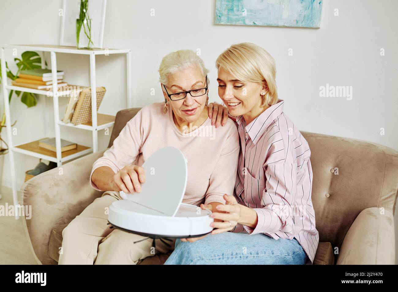 Ritratto di donna anziana con capelli bianchi che tiene l'aspirapolvere robot con una figlia giovane che mostra dispositivi smart home Foto Stock