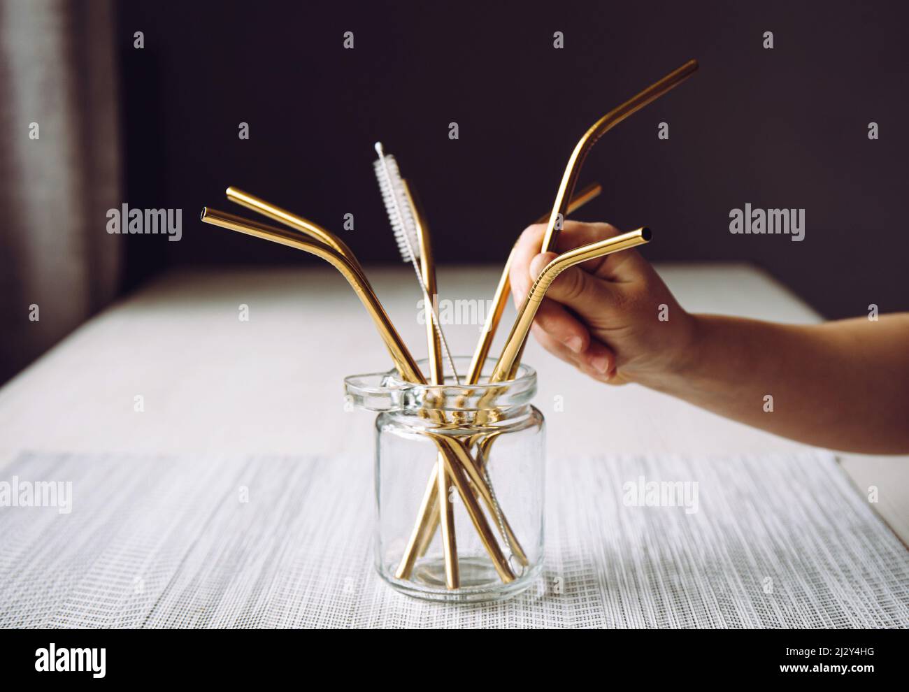 La mano del bambino prende la cannuccia bevente del metallo d'oro dal vaso di vetro nella cucina domestica. Concetto di stile di vita sostenibile. Foto Stock