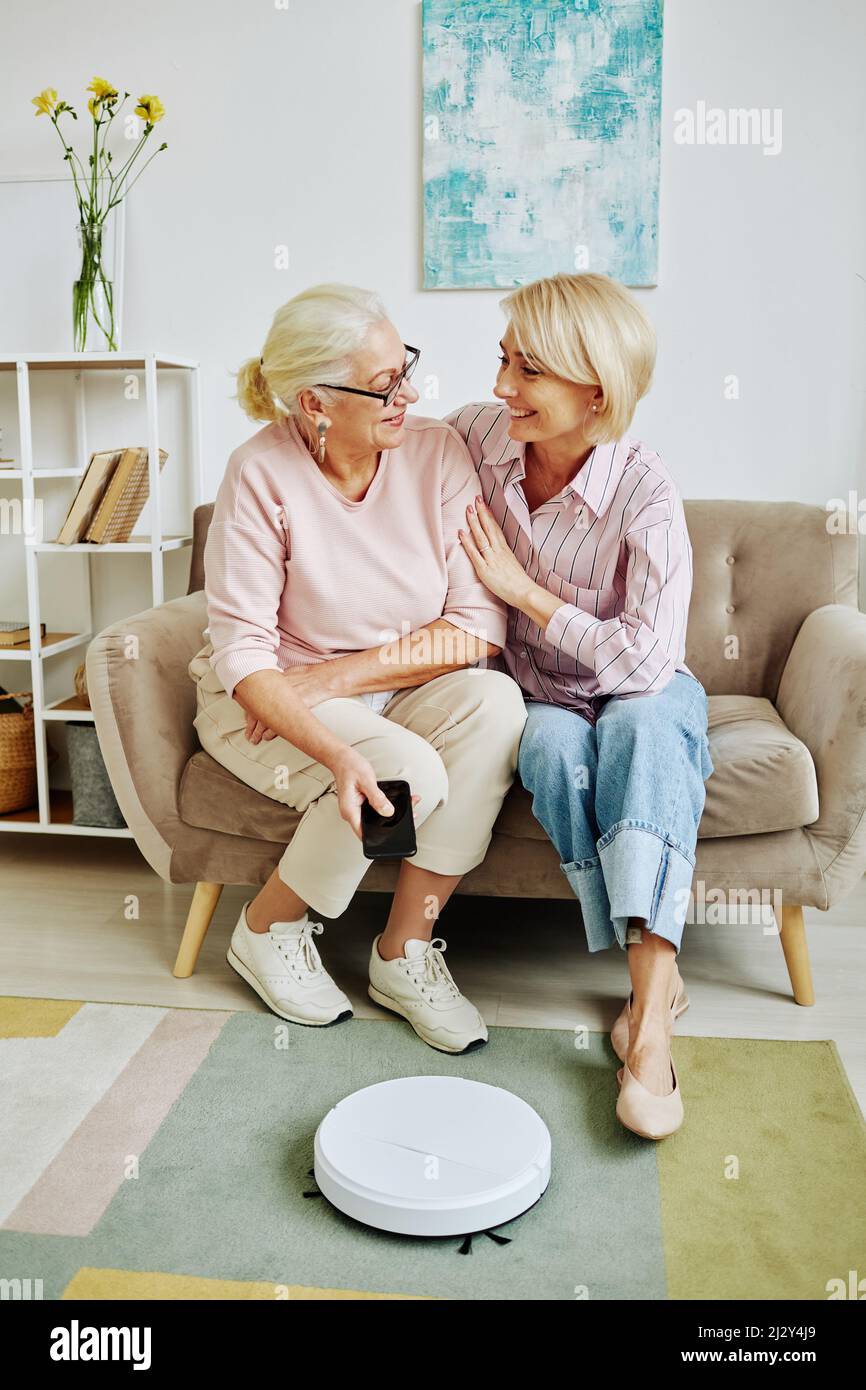 Ritratto verticale di una donna anziana sorridente che collega l'aspirapolvere robot allo smartphone durante la configurazione di dispositivi domestici intelligenti con il perno della figlia di un adulto Foto Stock