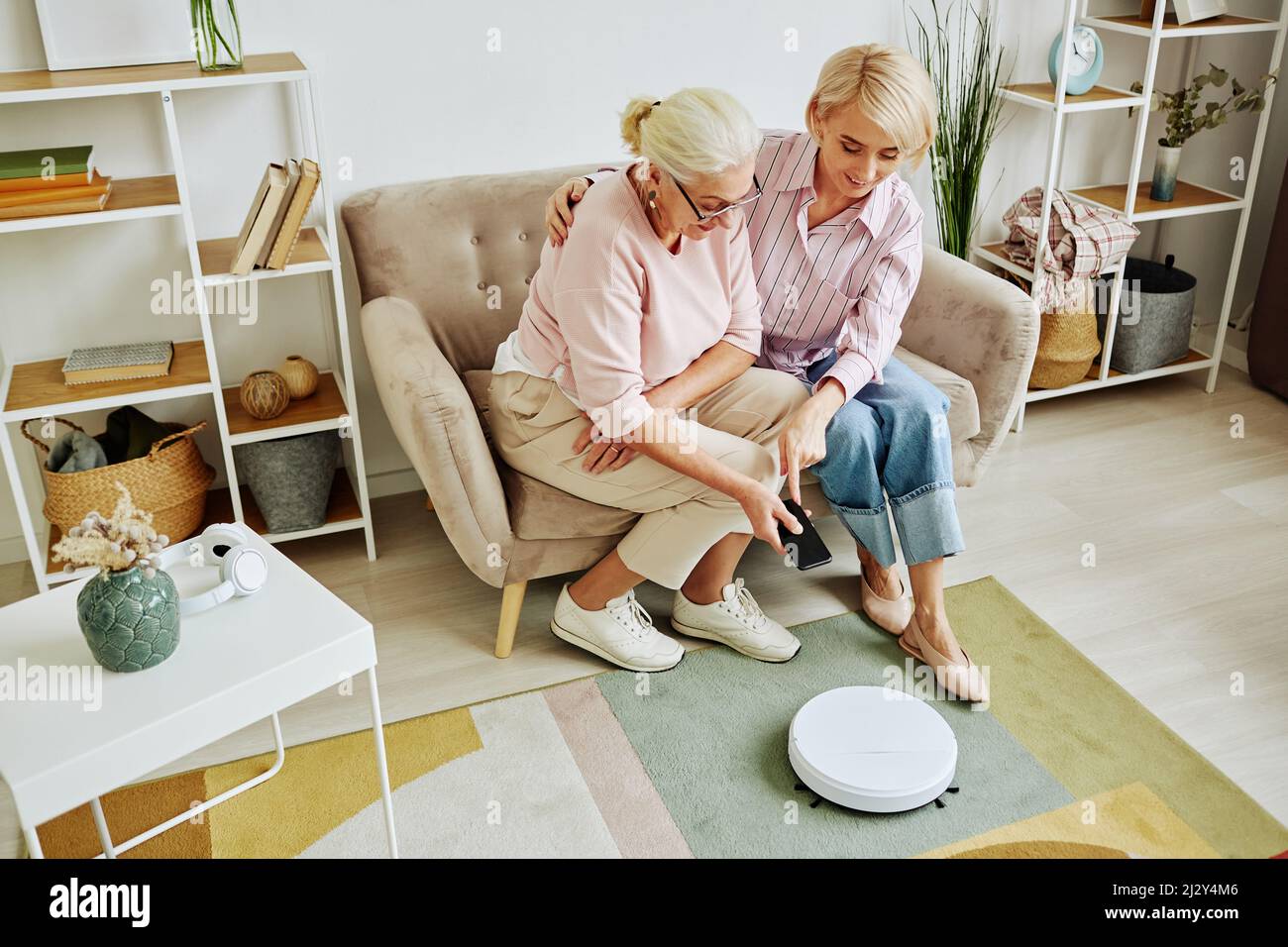 Ritratto ad angolo elevato di donna anziana che utilizza l'aspirapolvere robot tramite smartphone durante la configurazione di dispositivi domestici intelligenti con l'aiuto di una figlia adulta Foto Stock